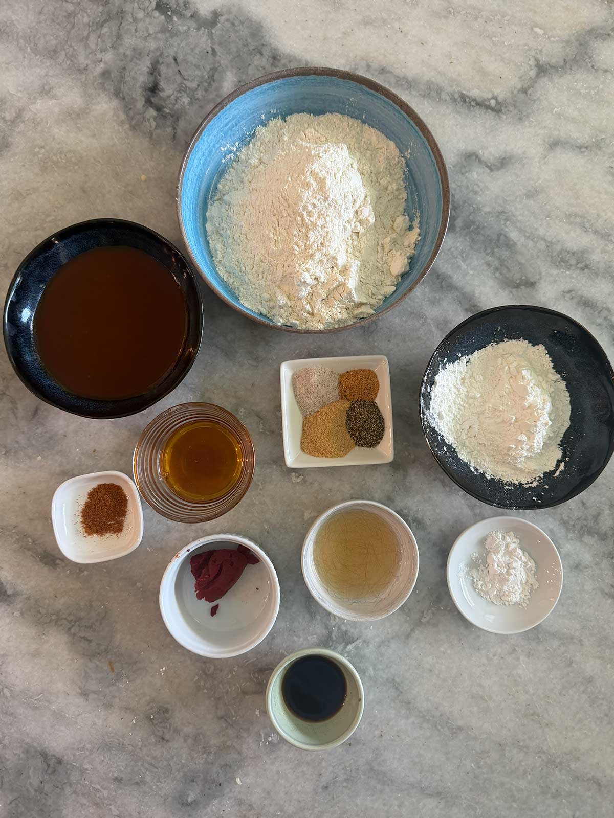 Top-down view of bowls containing flour, spices, oil, soy sauce, tomato paste, and other baking or cooking ingredients arranged on a marble countertop.