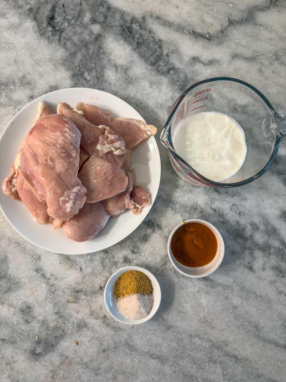 Raw chicken breasts on a white plate, with a measuring cup of milk, a small bowl of mustard, and a small bowl of mixed seasonings on a marble countertop.