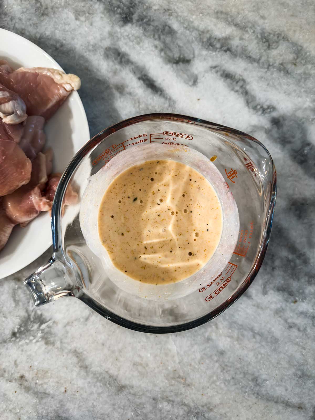 A glass measuring cup filled with a creamy, seasoned marinade sits on a gray countertop next to a plate of raw chicken pieces.