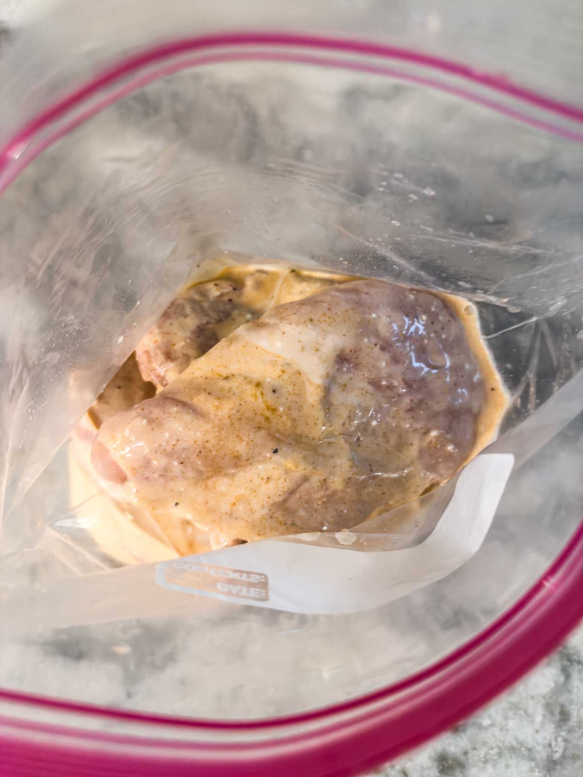 Raw chicken marinating in a zip-top plastic bag with a creamy, seasoned mixture, viewed from above. The bag is partially open and sitting on a countertop.