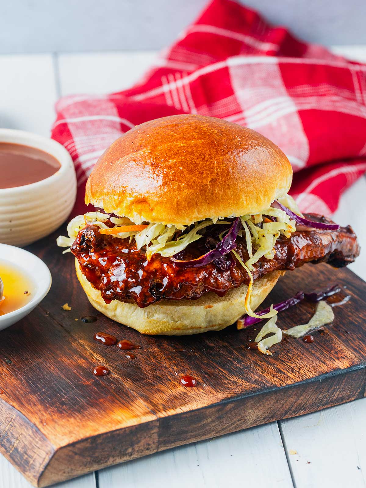 A honey barbecue chicken sandwich with slaw on a brioche bun sits on a wooden board, with a red checkered napkin and dipping sauces in the background.