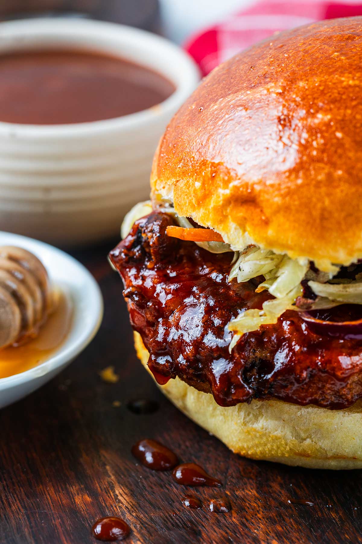 Close-up of a barbecue sauced chicken sandwich with shredded onions on a shiny bun, beside a bowl of dark sauce and a dish of honey with a honey dipper on a wooden surface.