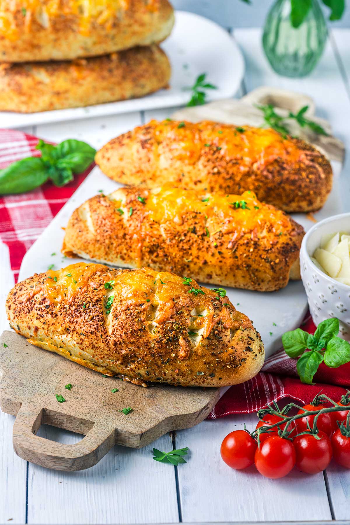 Three cheesy garlic bread loaves on a wooden board, garnished with herbs. A bowl of butter, basil leaves, and cherry tomatoes sit nearby. More bread loaves are in the background on a white plate.