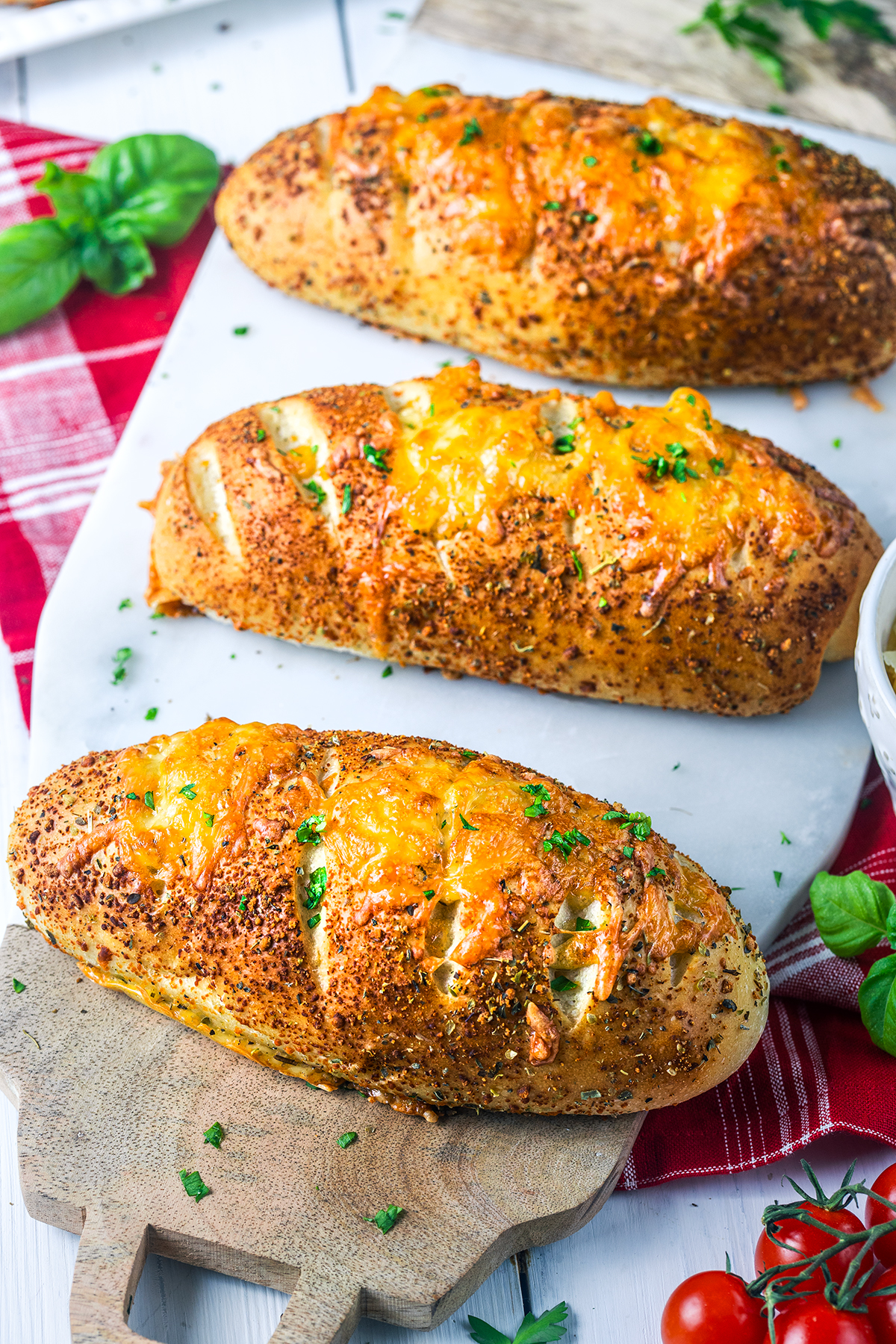 Three cheesy garlic bread loaves topped with melted cheese and herbs sit on a marble board, surrounded by basil leaves, cherry tomatoes, and a red checkered napkin.