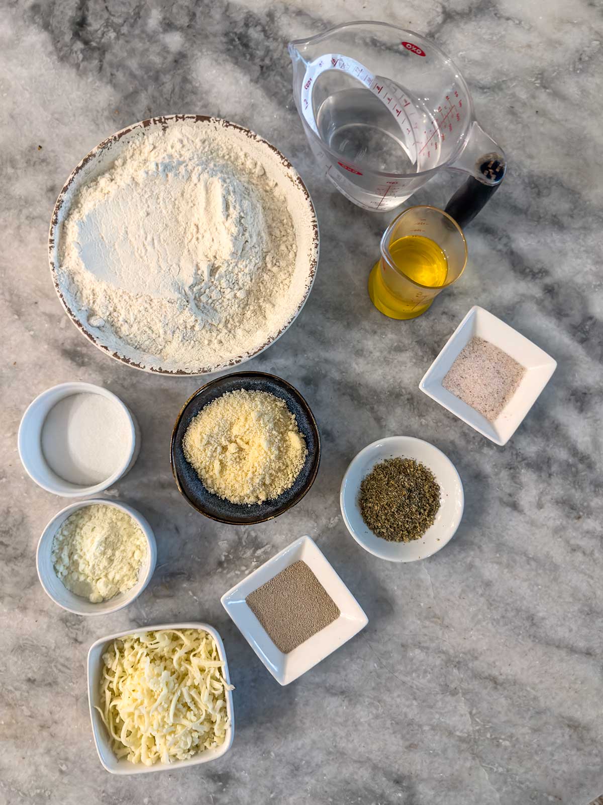 Assorted bread dough and topping ingredients on a marble counter, including flour, water, oil, grated cheese, yeast, herbs, salt, and sugar in various bowls and containers.