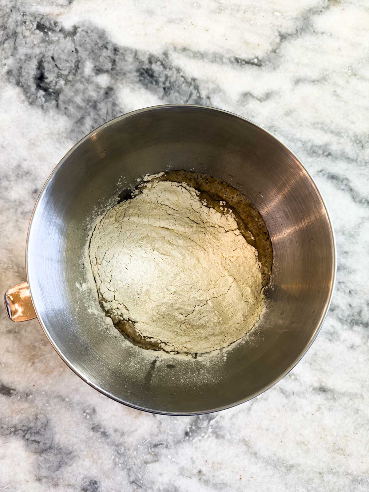 A metal mixing bowl with flour and other baking ingredients is sitting on a marble countertop, viewed from above.