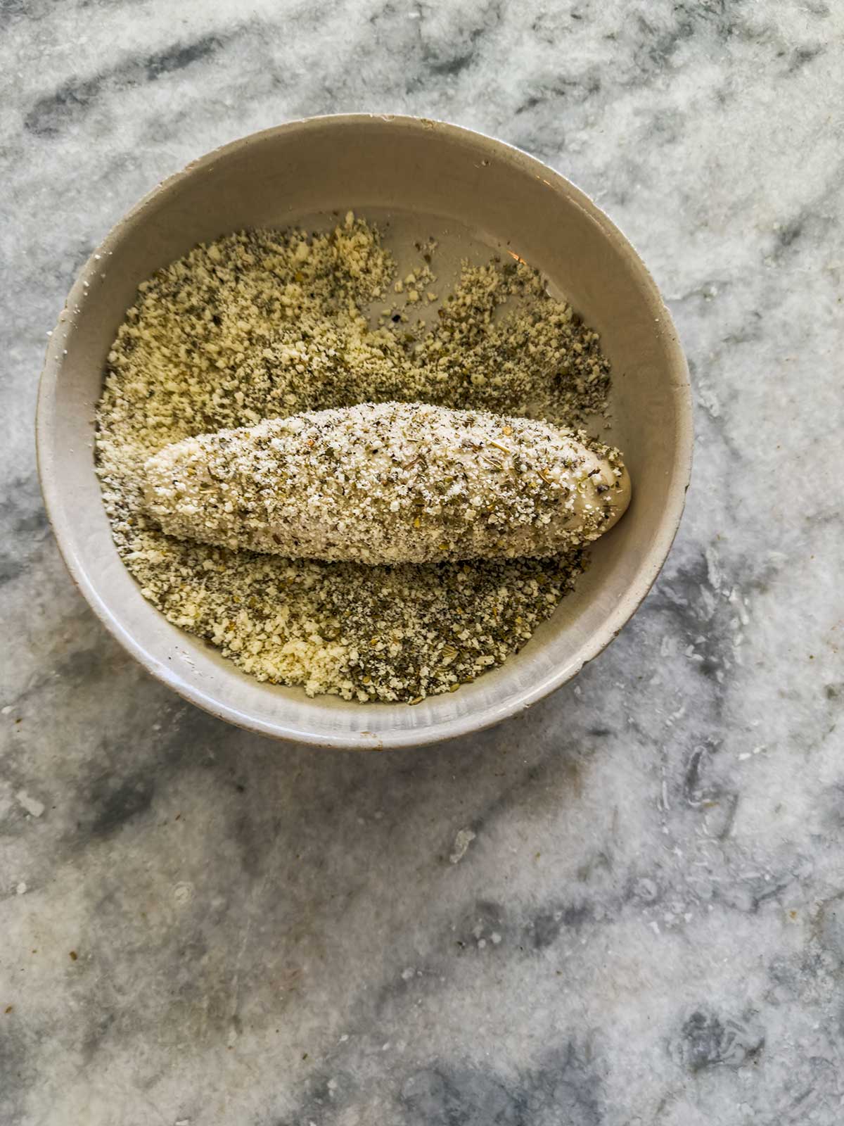 A small bowl containing a coated piece of food, possibly cheese or tofu, being rolled in a mixture of herbs or breadcrumbs on a gray marble surface.