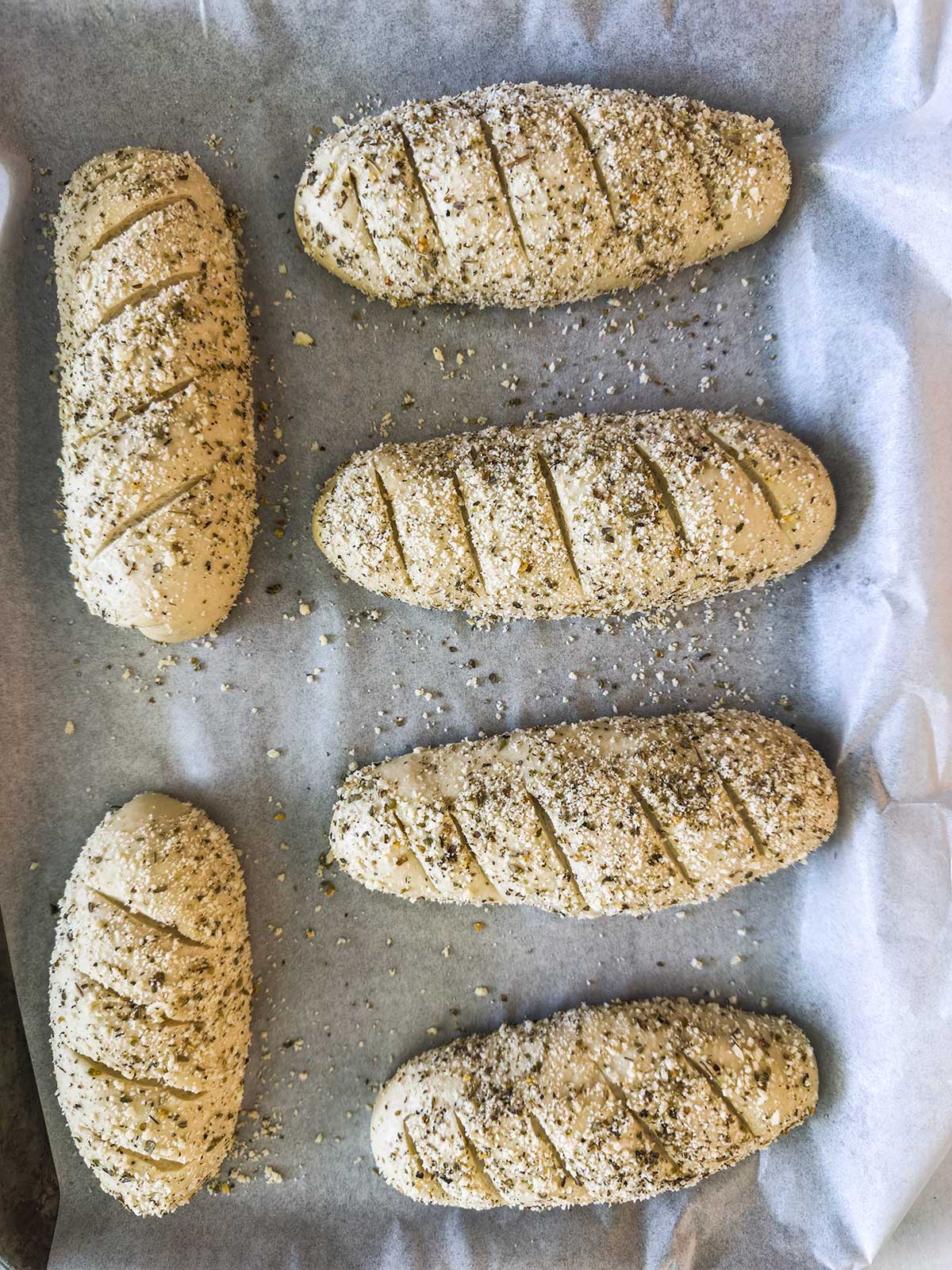 Six unbaked loaves of seeded bread dough with diagonal slashes on top rest on a parchment-lined baking sheet, ready to go into the oven.