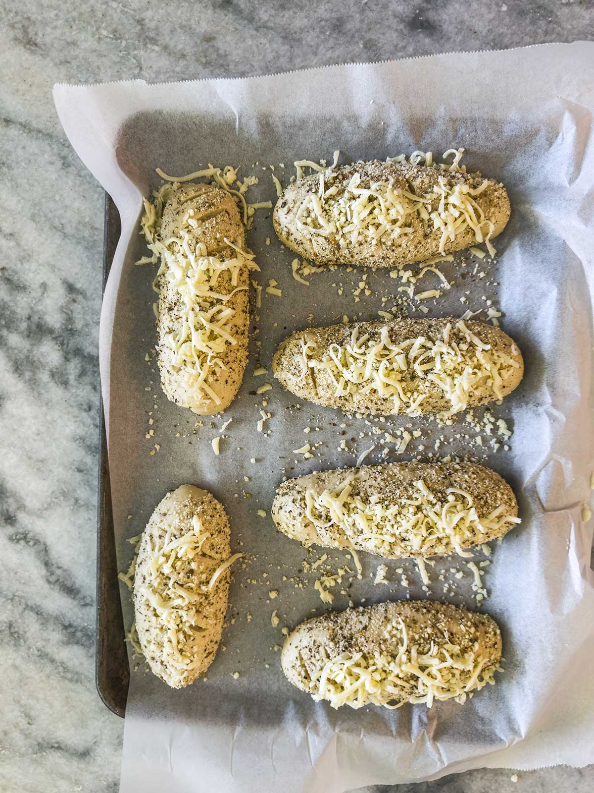 Six unbaked bread rolls topped with shredded cheese and seeds on a parchment-lined baking sheet, placed on a gray marble surface.