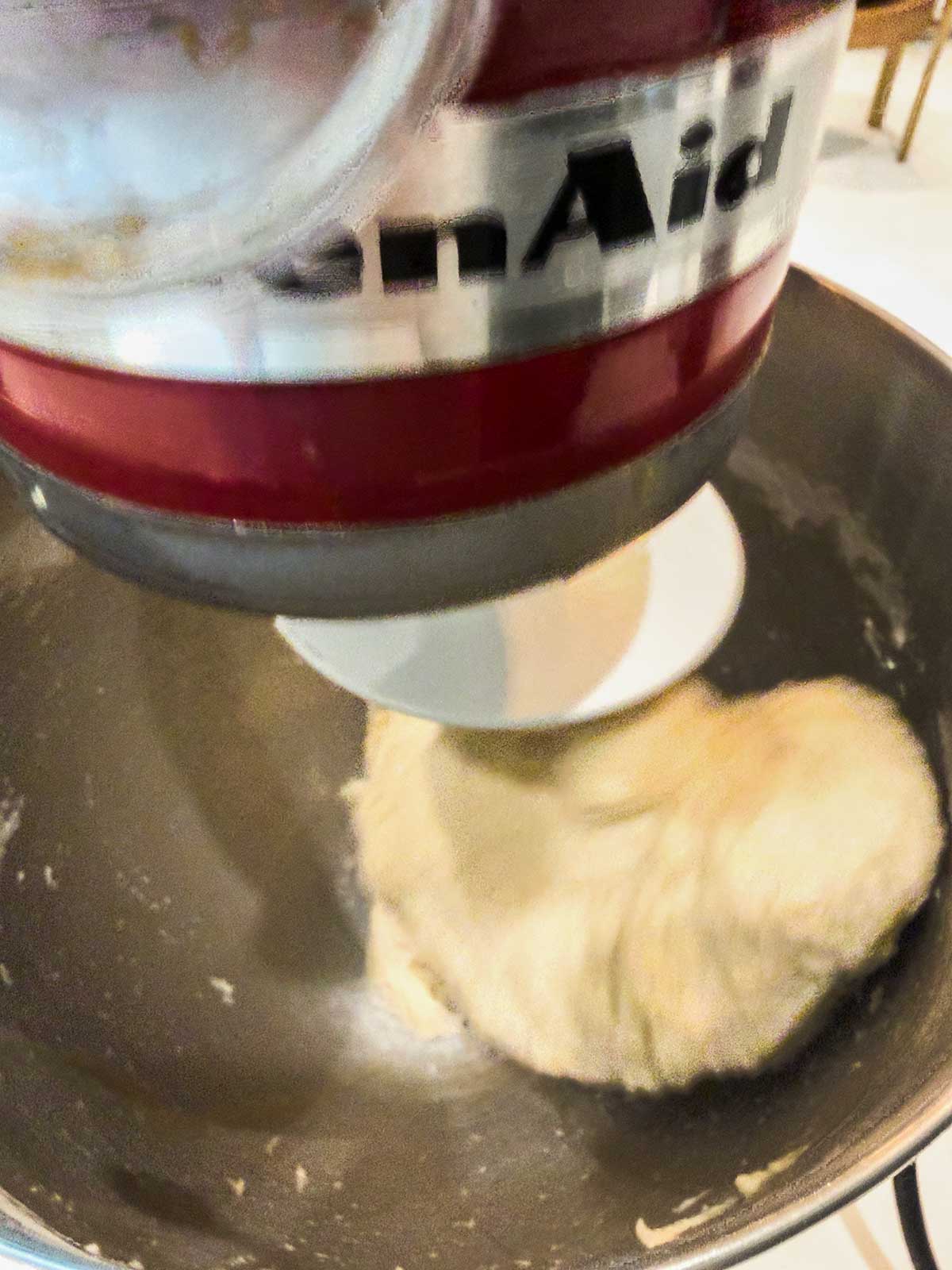 A close-up of dough being kneaded inside a mixing bowl by a red KitchenAid stand mixer with a dough hook attachment.