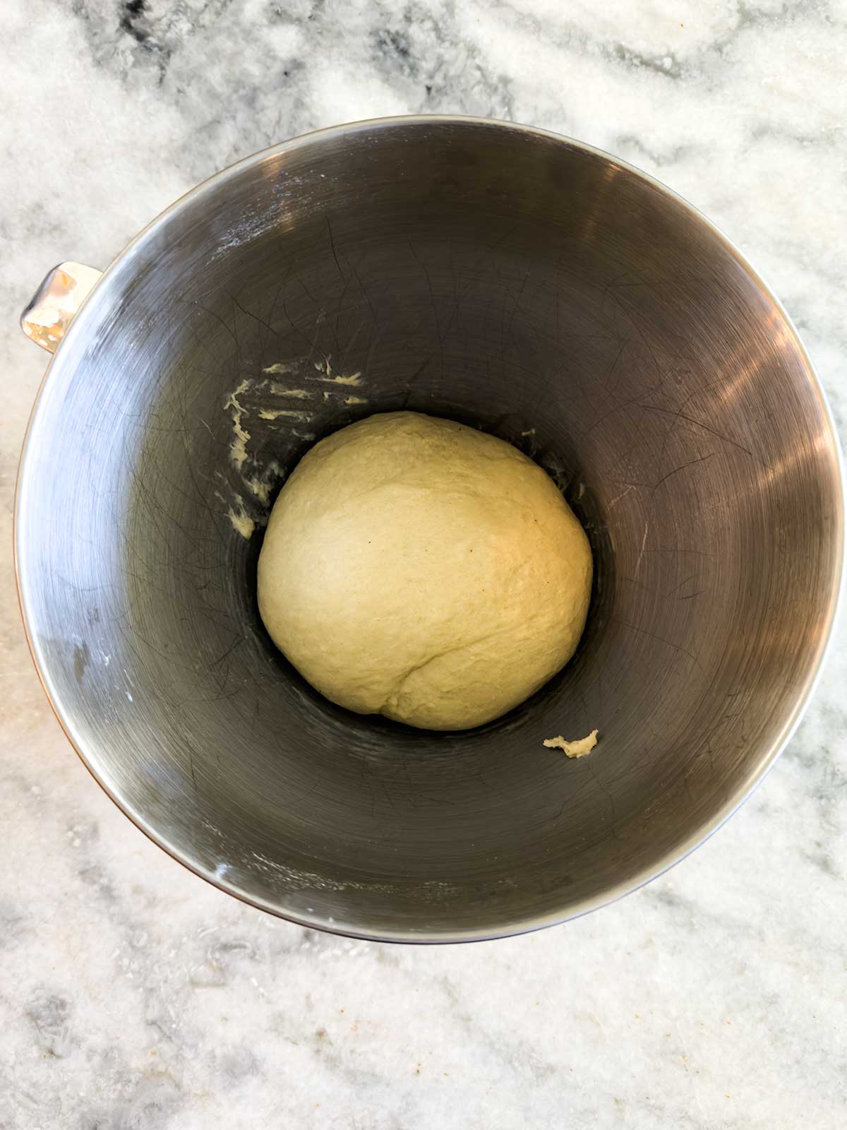 A ball of dough sits in the center of a metal mixing bowl on a marble countertop.