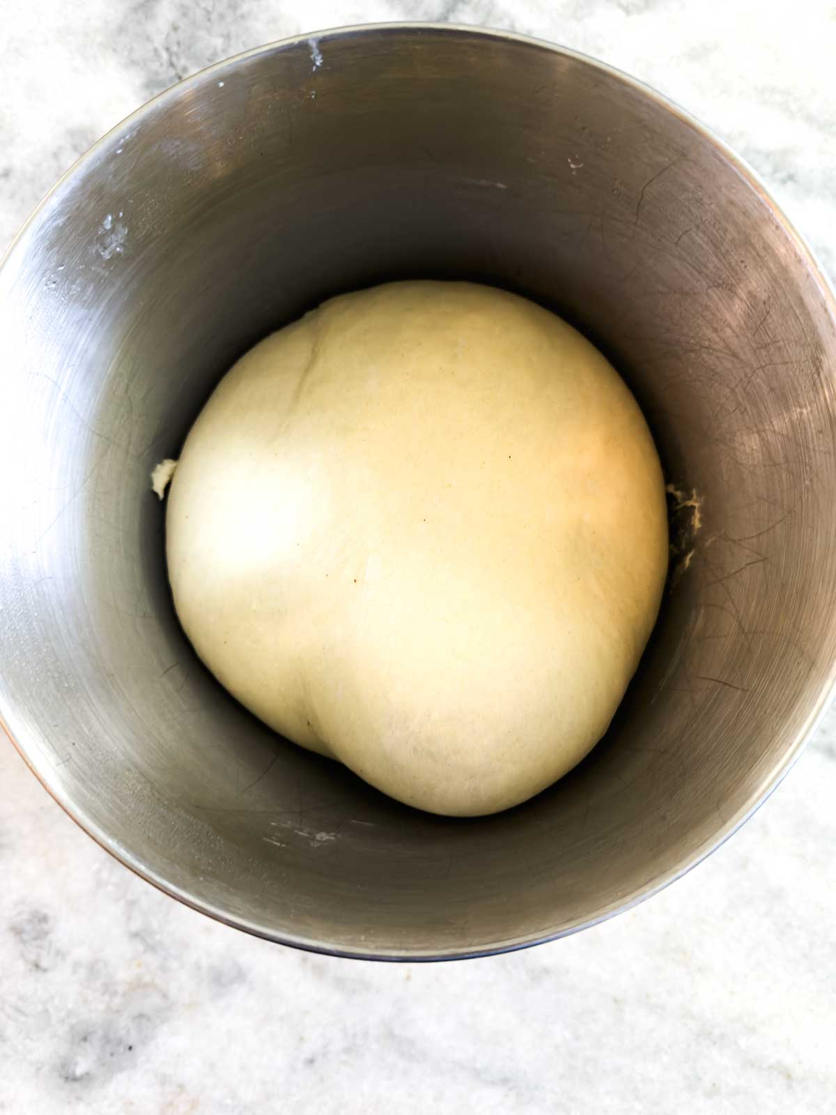 A smooth ball of dough rests in a large metal mixing bowl on a light-colored countertop, ready for baking or further preparation.