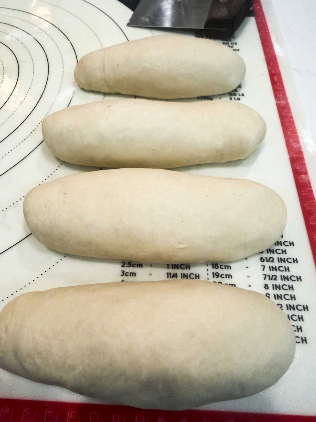 Four evenly shaped, uncooked bread dough logs are lined up on a baking mat with measurement markings.