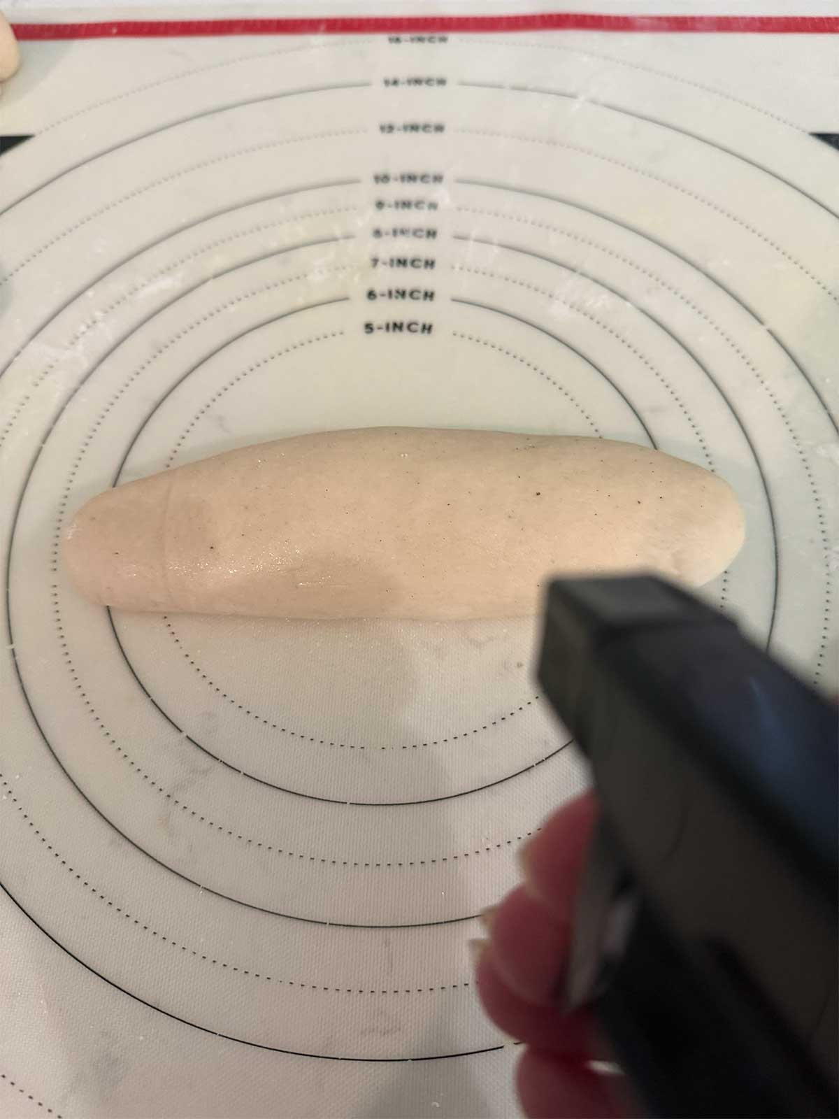A hand sprays water onto a shaped dough log resting on a silicone mat with circular measurement guides marked in inches.