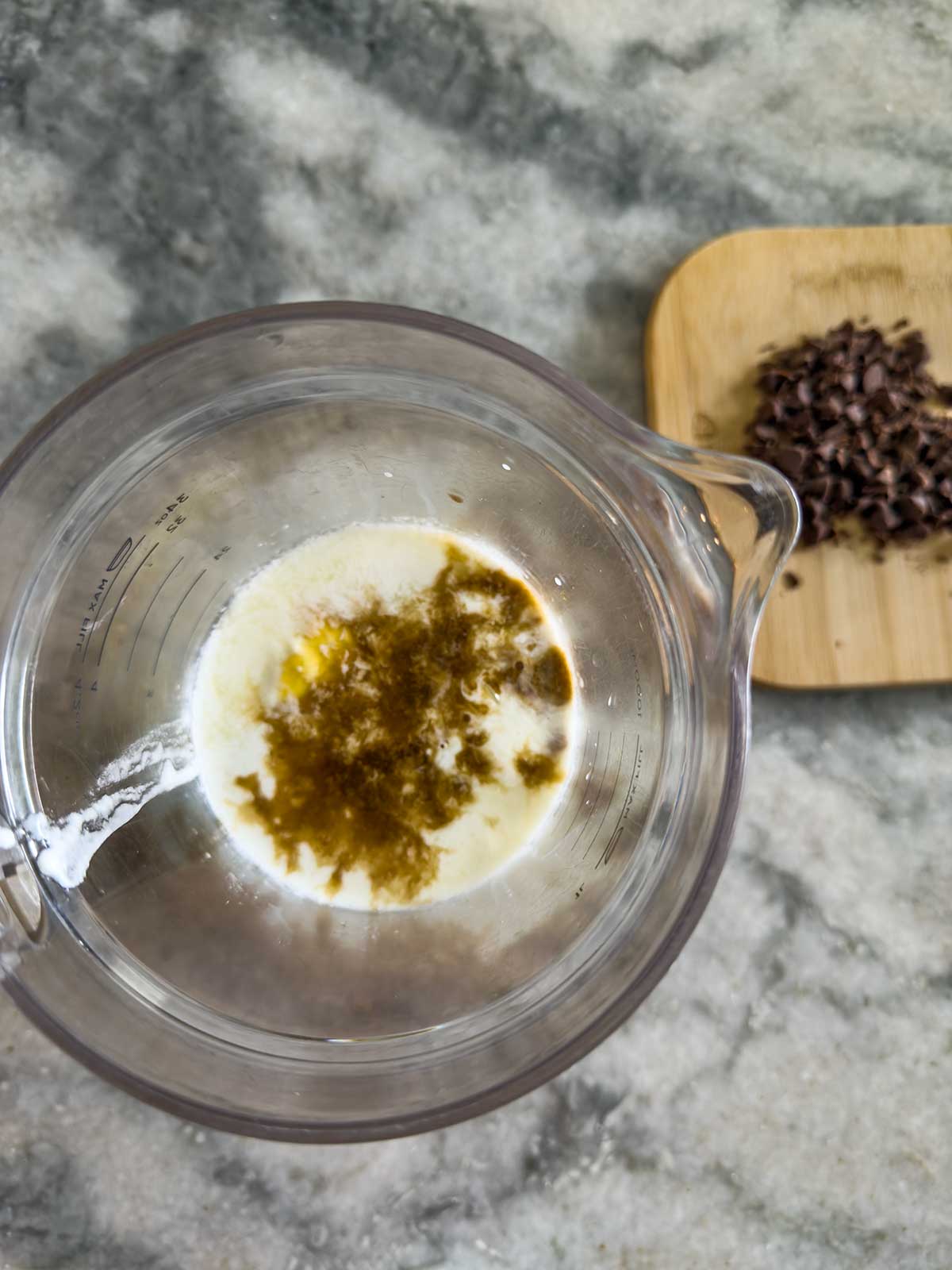 A clear liquid with a brown swirl rests in a measuring cup, hinting at a ninja creami frozen yogurt mixture. Nearby, on a wooden board, lies a tempting pile of chocolate chips.