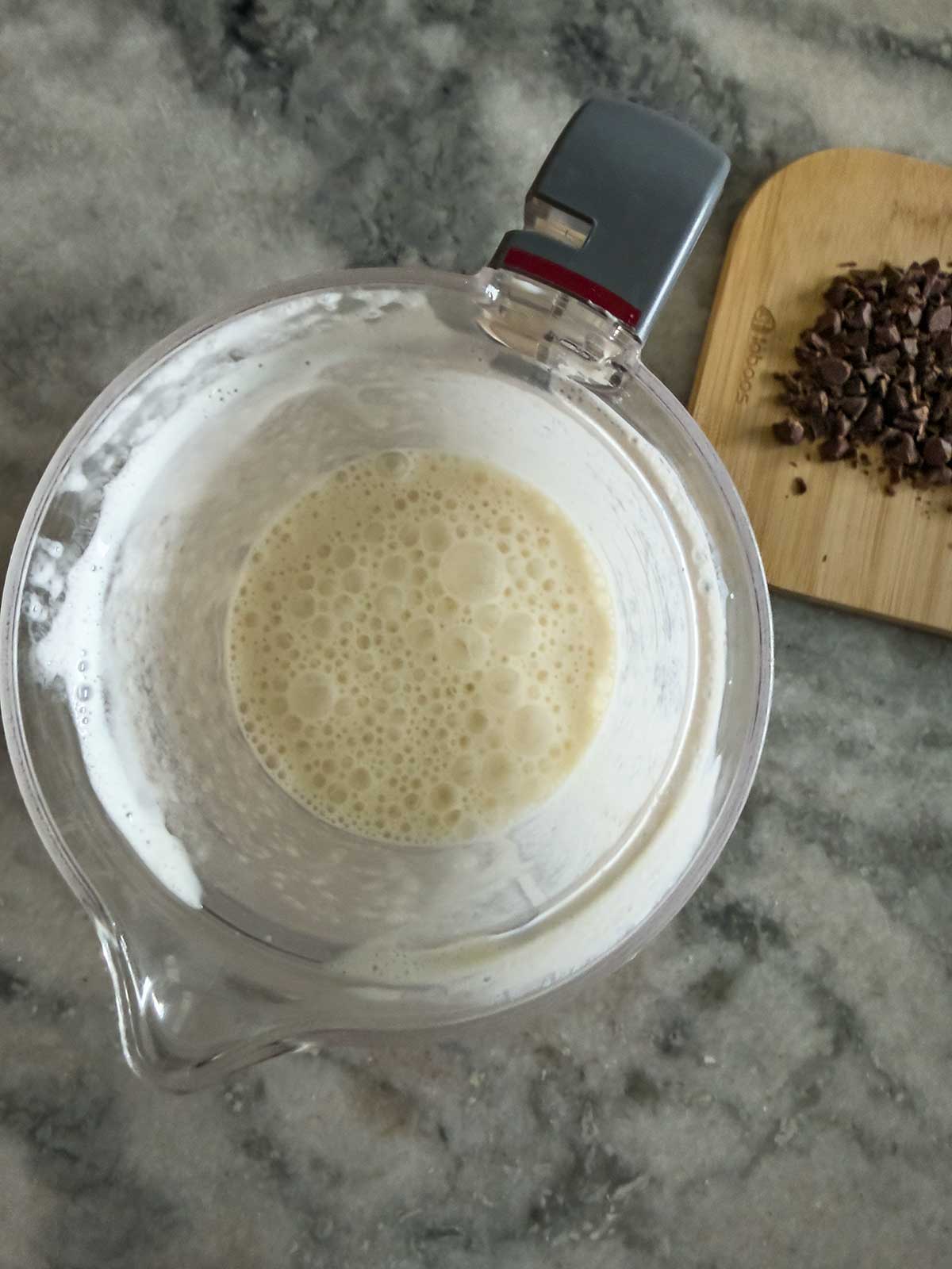 A blender pitcher filled with a frothy, creamy liquid—reminiscent of Ninja Creami frozen yogurt—sits on a marble counter next to a wooden cutting board with chopped chocolate pieces.