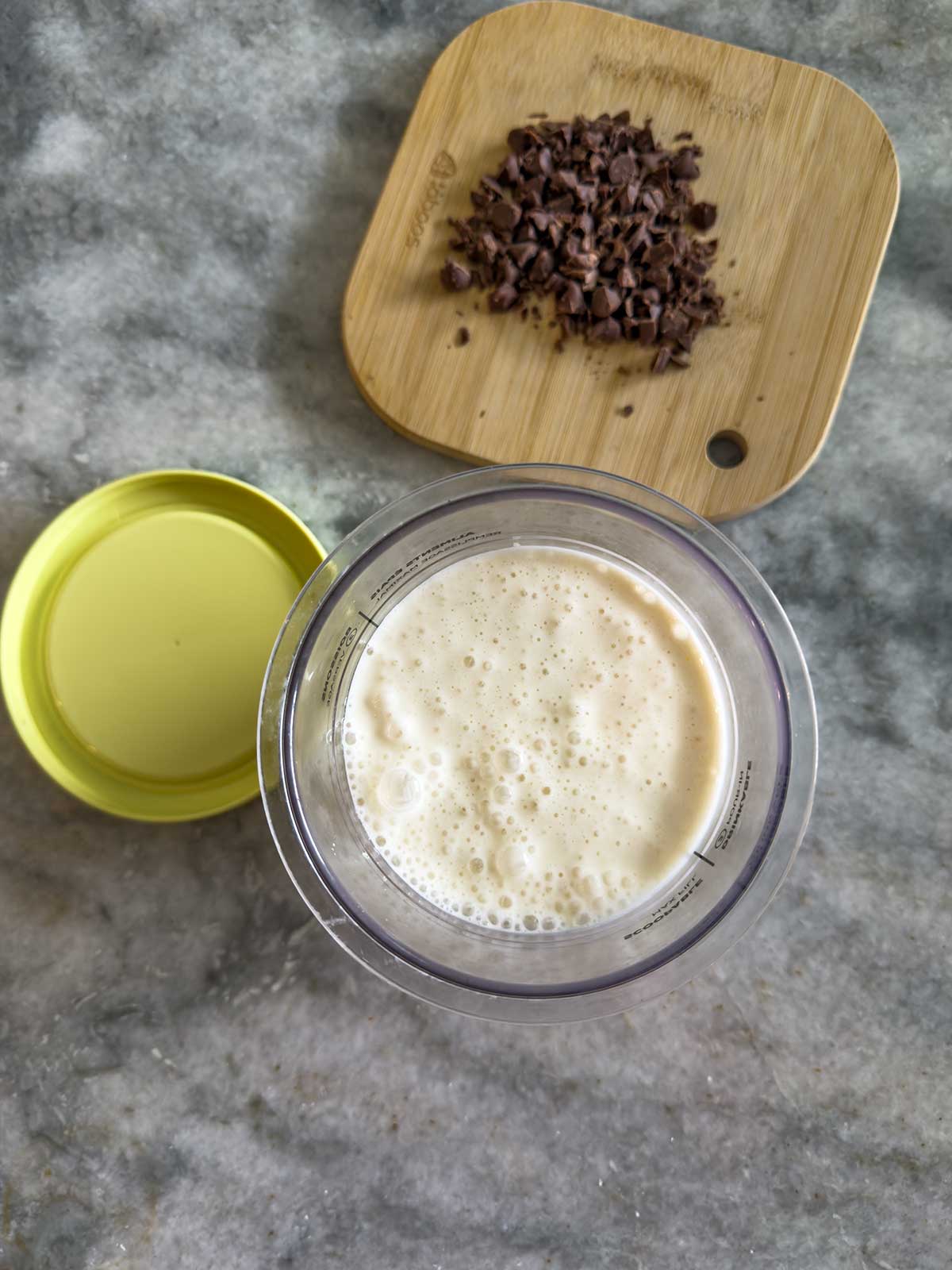 A ninja creami pint cup filled with a frothy light-colored smoothie, made using the Ninja Creami frozen yogurt method, sits on a gray counter. Behind it is a wooden board with chopped chocolate pieces and a yellow lid resting nearby.