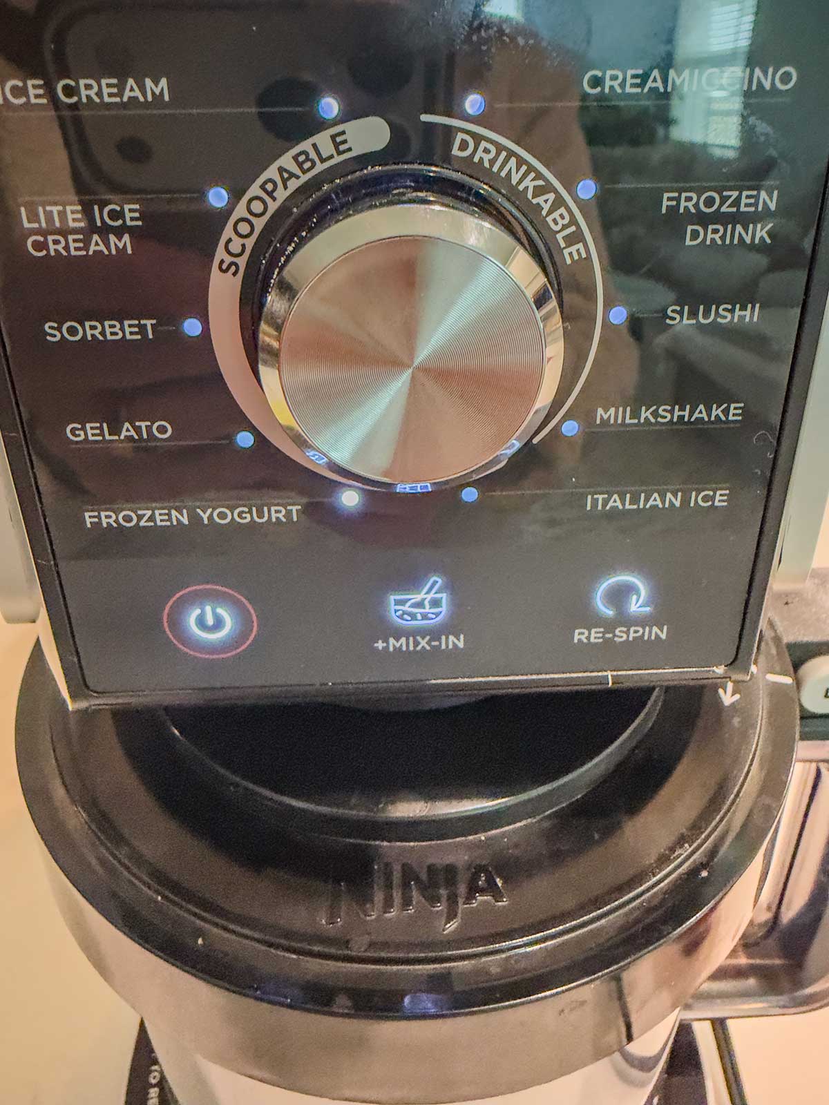 Close-up of a Ninja Creami t maker control panel with a central dial labeled Scoopable and Drinkable. Options include ice cream, sorbet, ninja creami frozen yogurt, gelato, milkshake, slushi, and others. Power and more below.