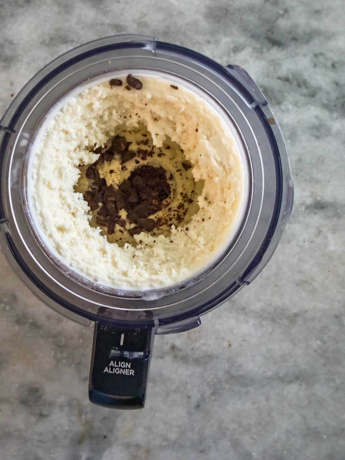 A Ninja Creami frozen yogurt mixture sits in a food processor bowl with chocolate pieces in the center, viewed from above on a gray marble countertop.