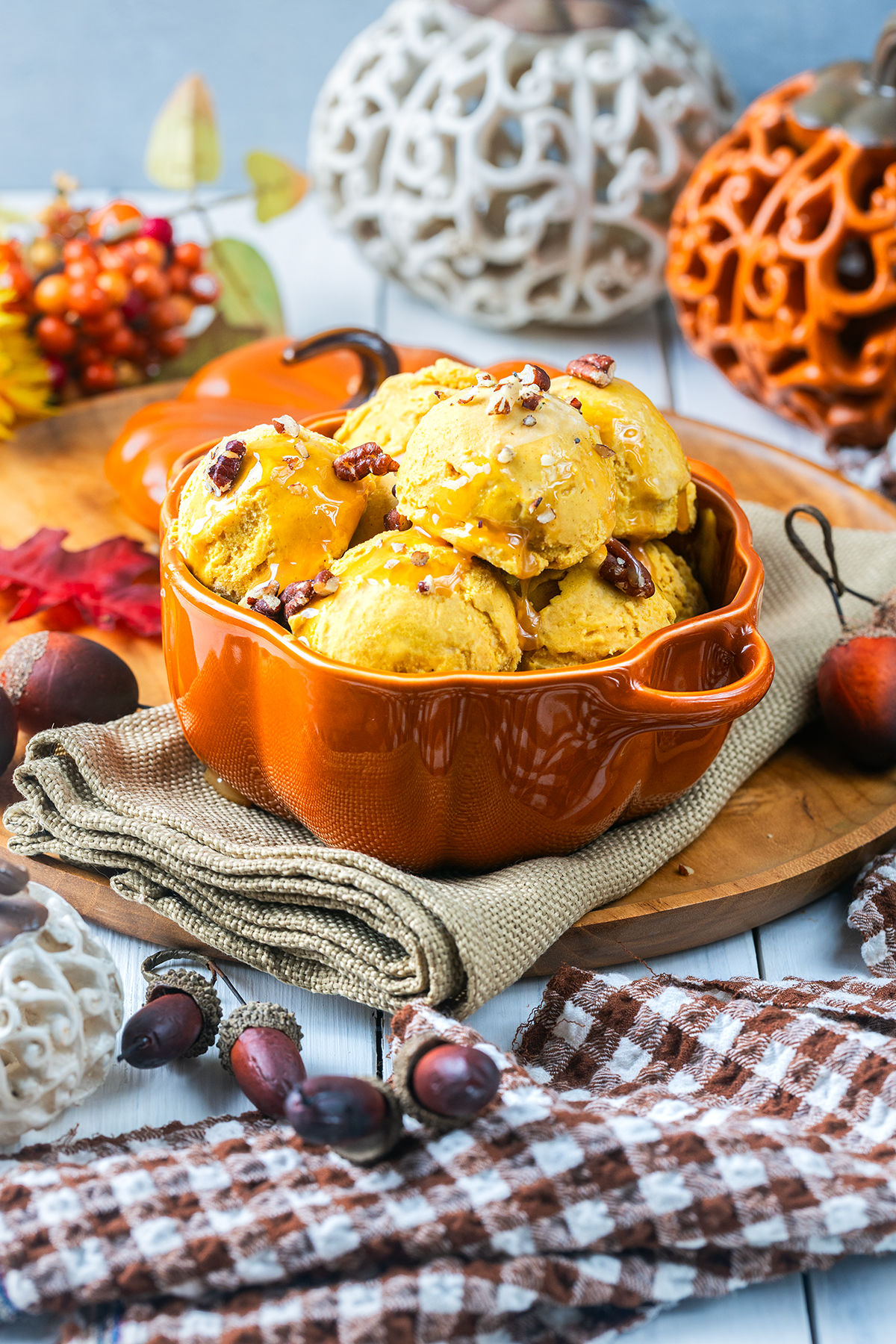 Pumpkin ice cream topped with pecans and caramel sauce in an orange pumpkin-shaped dish, surrounded by autumn decor, chestnuts, and a checkered cloth on a rustic wooden table.