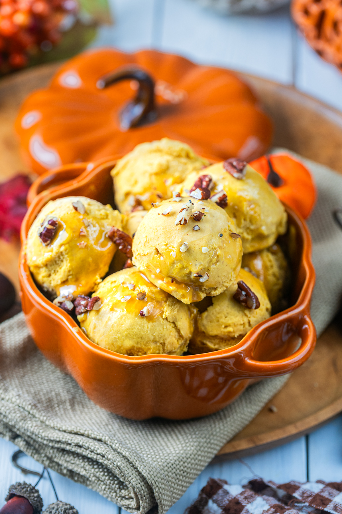 Pumpkin ice cream scoops in a pumpkin-shaped orange bowl, topped with nuts and syrup, sit on a beige cloth. Autumn-themed decor is in the background, including a ceramic pumpkin and fall leaves.