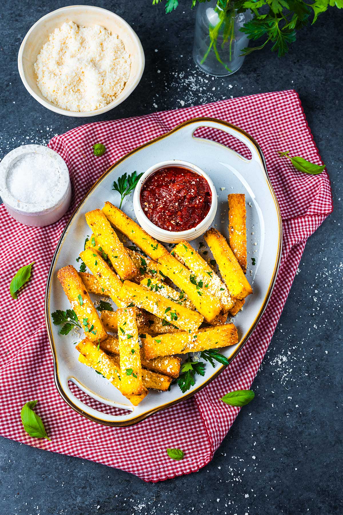 Golden polenta fries topped with grated cheese and herbs on a white platter, served with marinara sauce. A bowl of grated cheese, salt, and fresh basil are nearby, all set on a red checkered cloth.