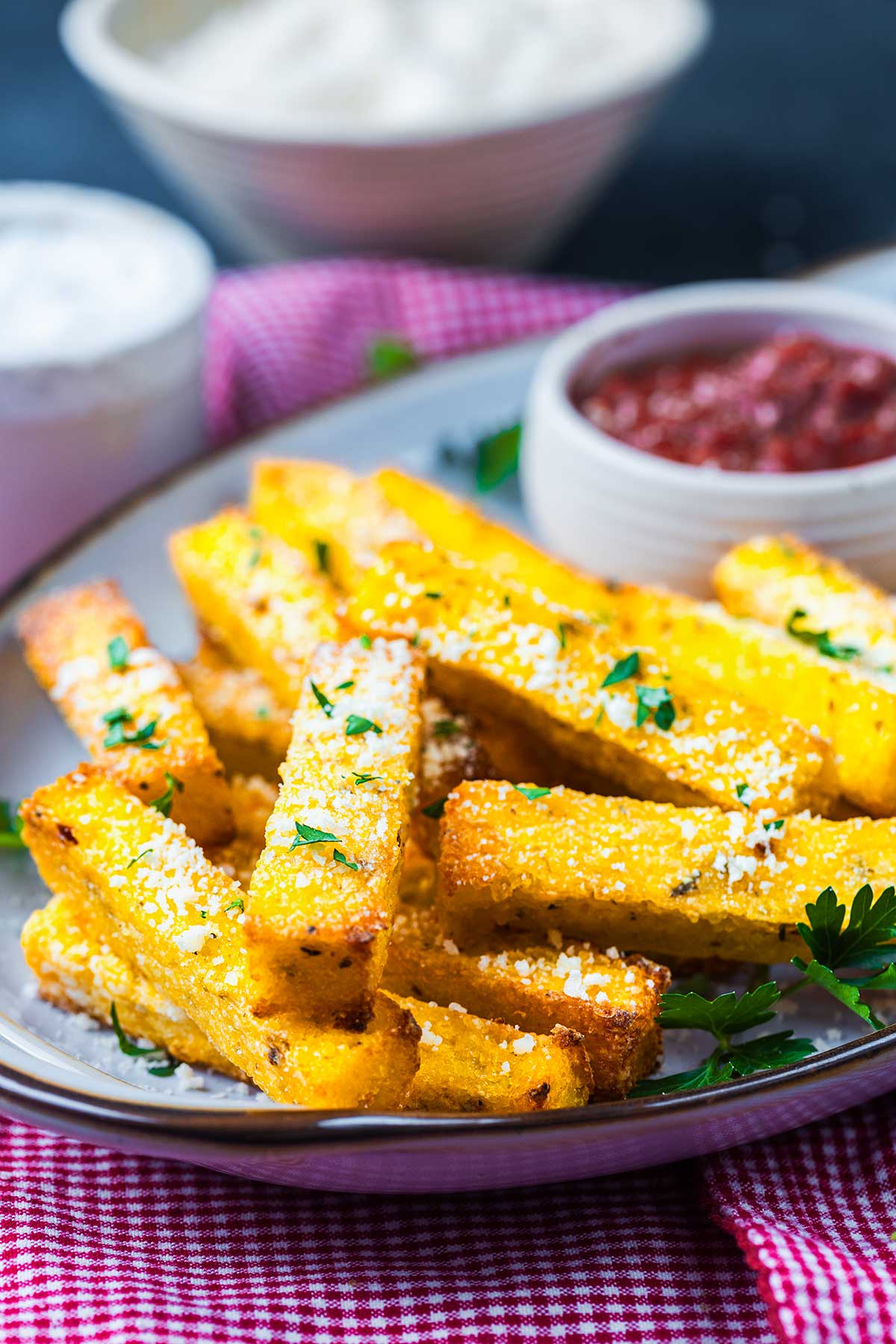 Golden polenta fries garnished with herbs and grated cheese, served on a plate with a dish of red dipping sauce and a blurred bowl in the background, set on a red checkered cloth.