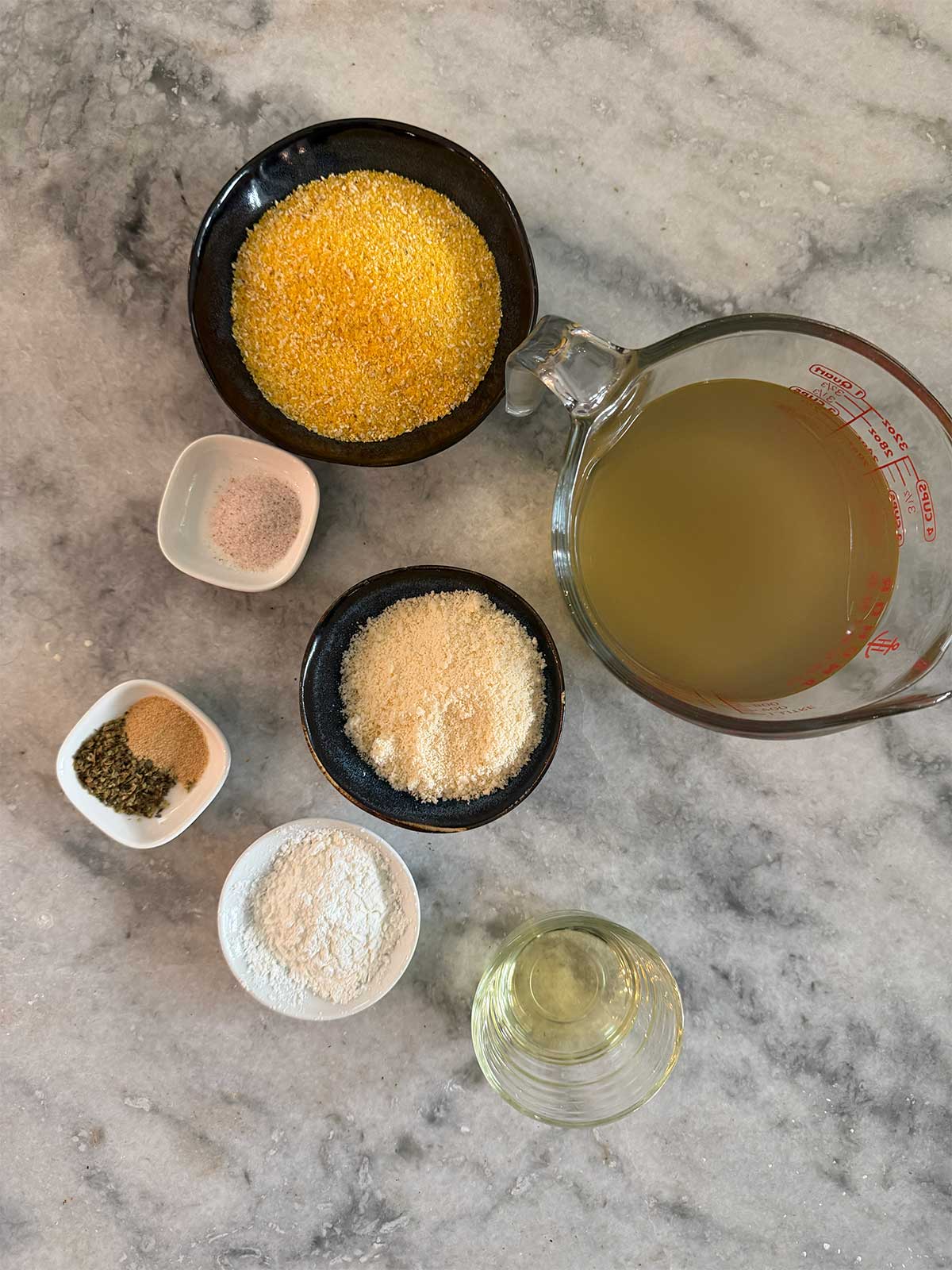 Bowls and cups holding polenta, broth, grated cheese, flour, oil, pepper, and salt are arranged on a gray marble surface, viewed from above.