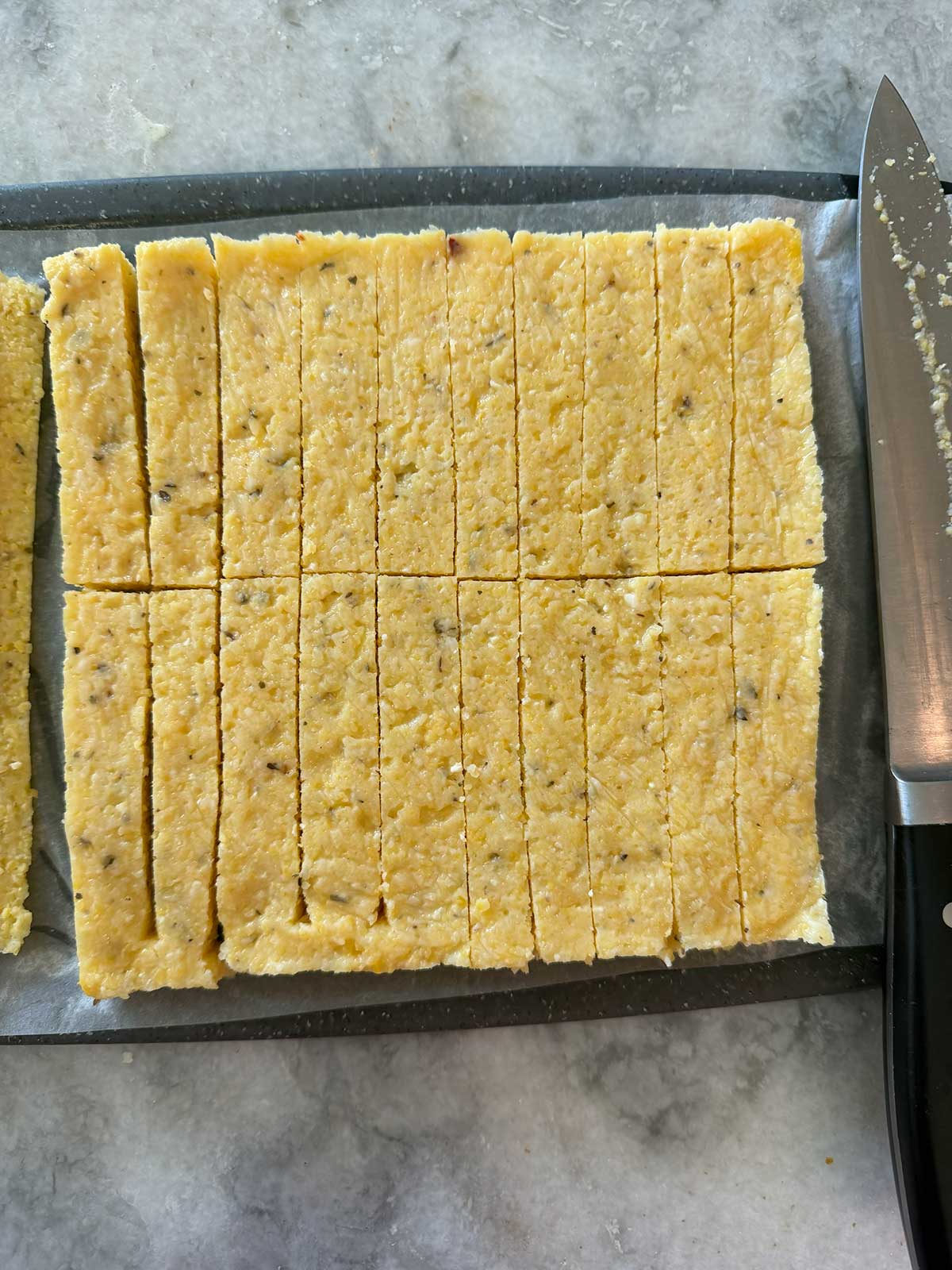 A rectangular slab of cornbread cut into even rectangular pieces sits on a tray. A knife with a black handle is placed on the right side, and the surface beneath is a light-colored countertop.