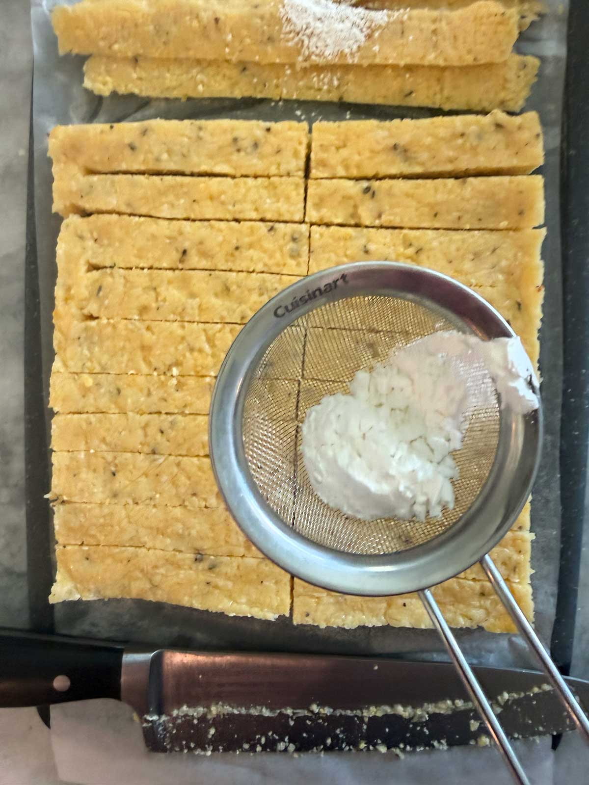 Unbaked rectangular polenta fries on parchment, a knife below, and a metal sifter with cornstarch resting on top of the dough.