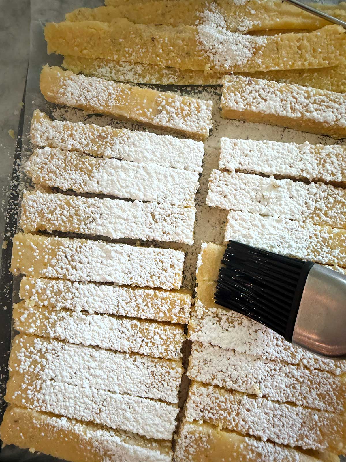 Rectangular polenta fries dusted with cornstarch are being brushed with a pastry brush with oil on it.