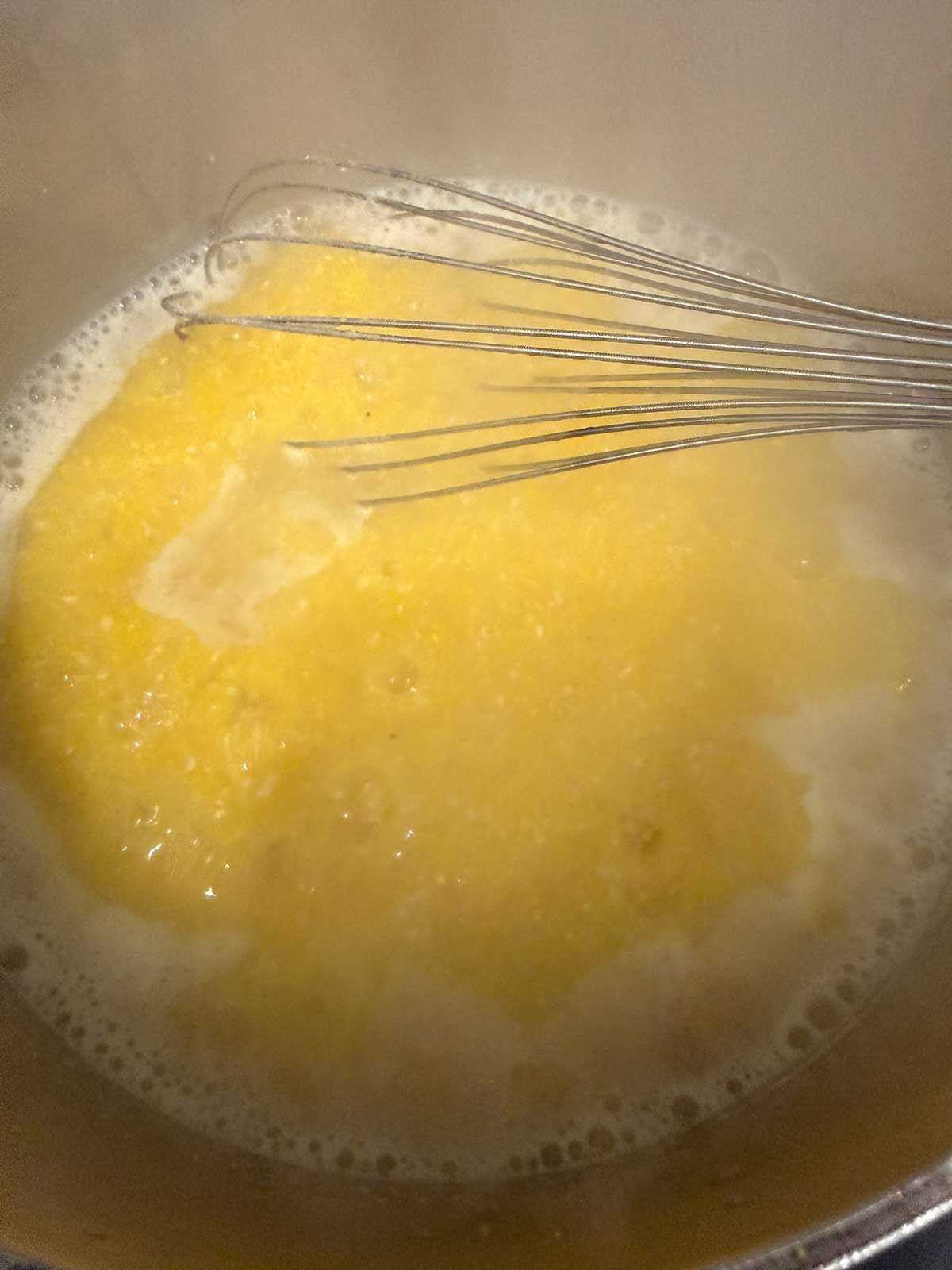 A metal whisk stirs yellow cornmeal in a pot of boiling water, making polenta. Steam rises from the mixture as it cooks.
