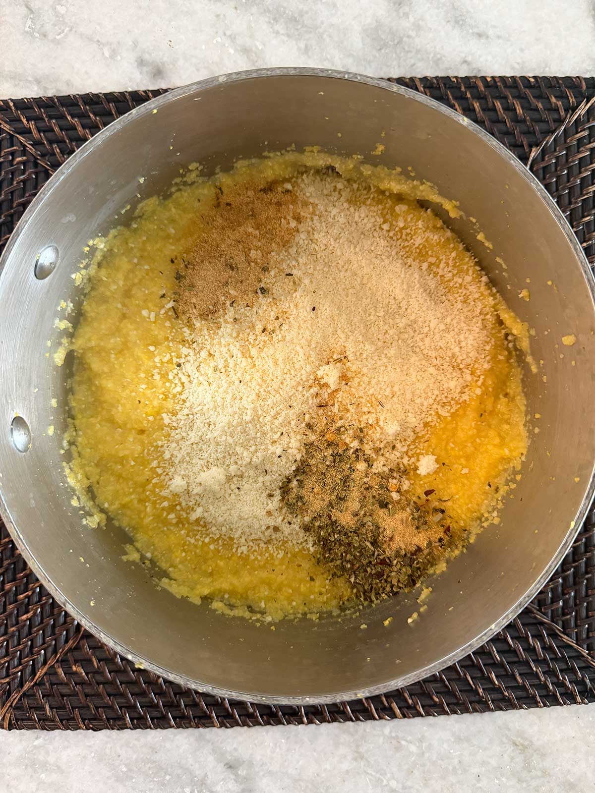 A pot of yellow polenta topped with grated cheese, breadcrumbs, and dried herbs sits on a woven placemat over a marble surface.