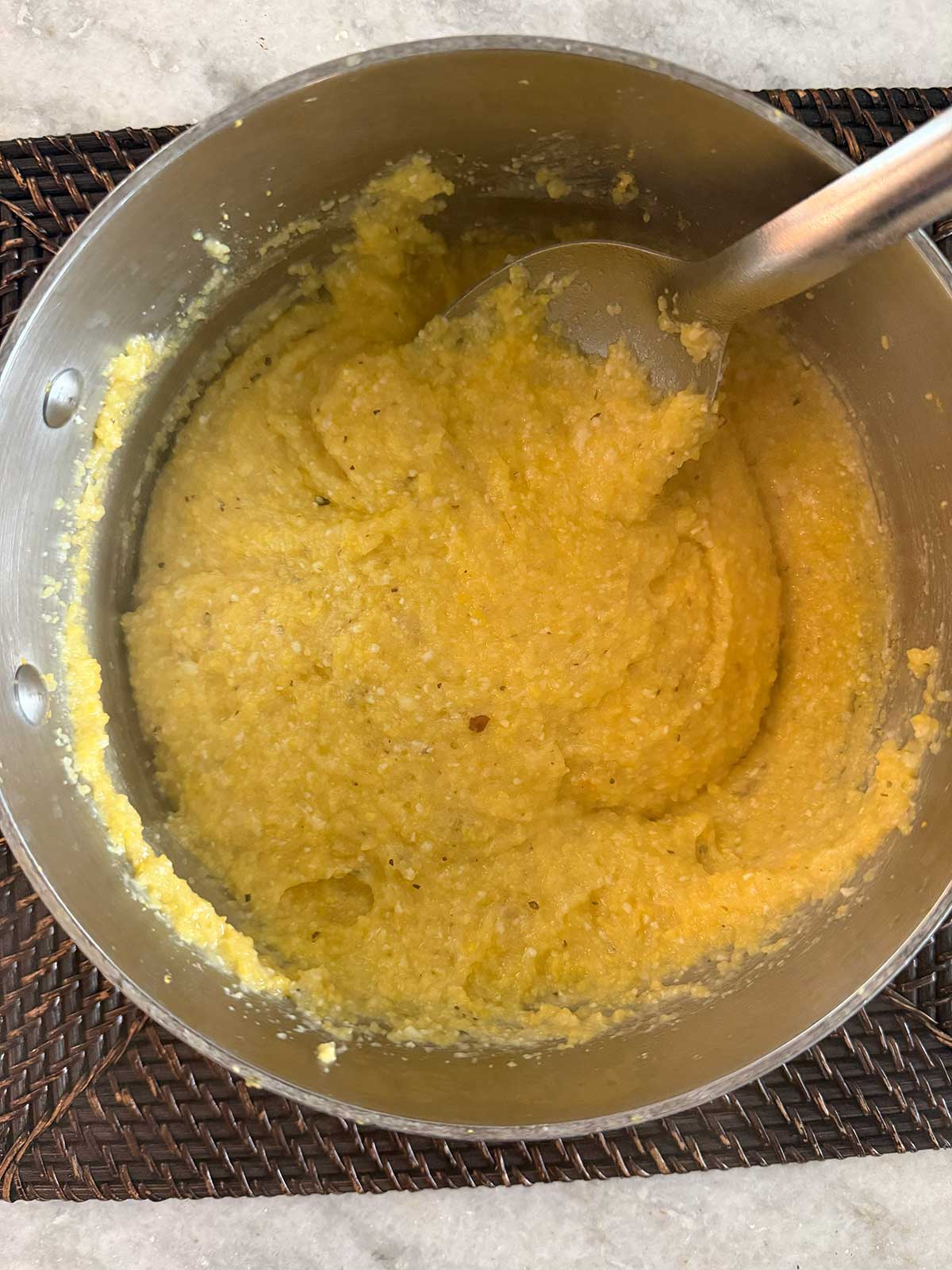 A pot of creamy, yellow cornmeal polenta being stirred with a metal spoon, sitting on a woven placemat.