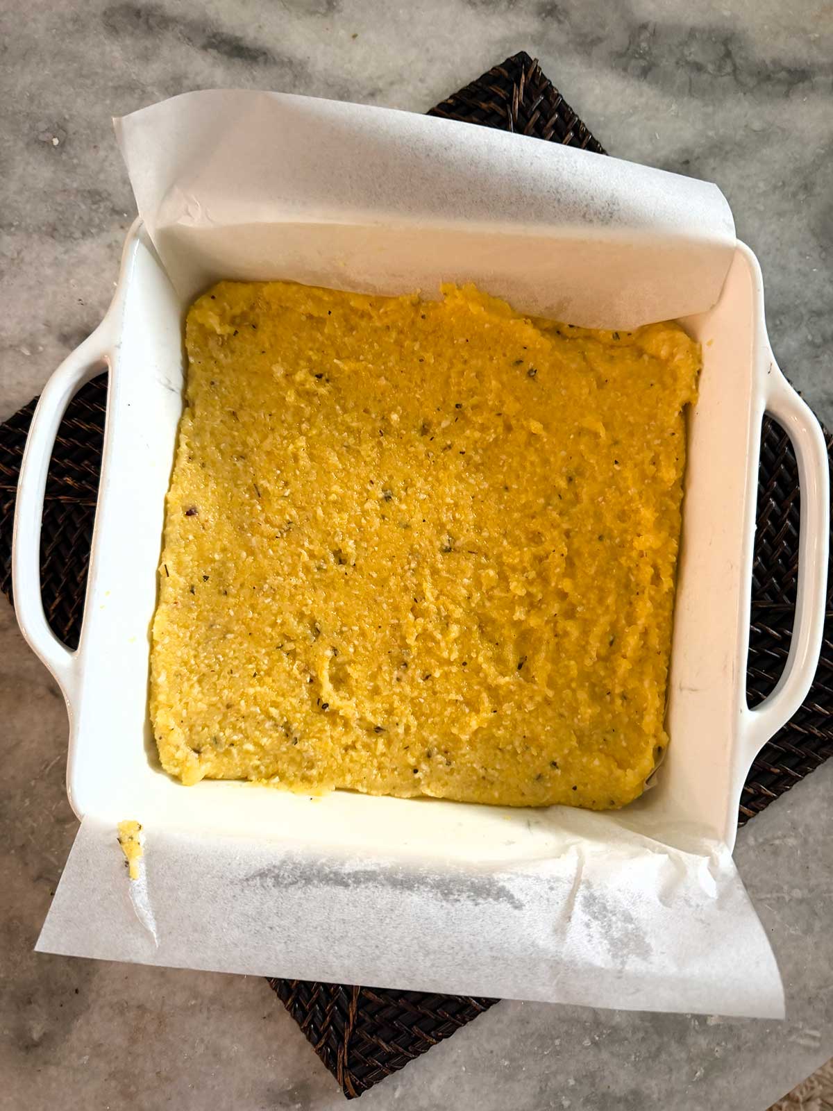 A white baking dish lined with parchment paper holds polenta, sitting on a woven dark mat on a marble countertop.