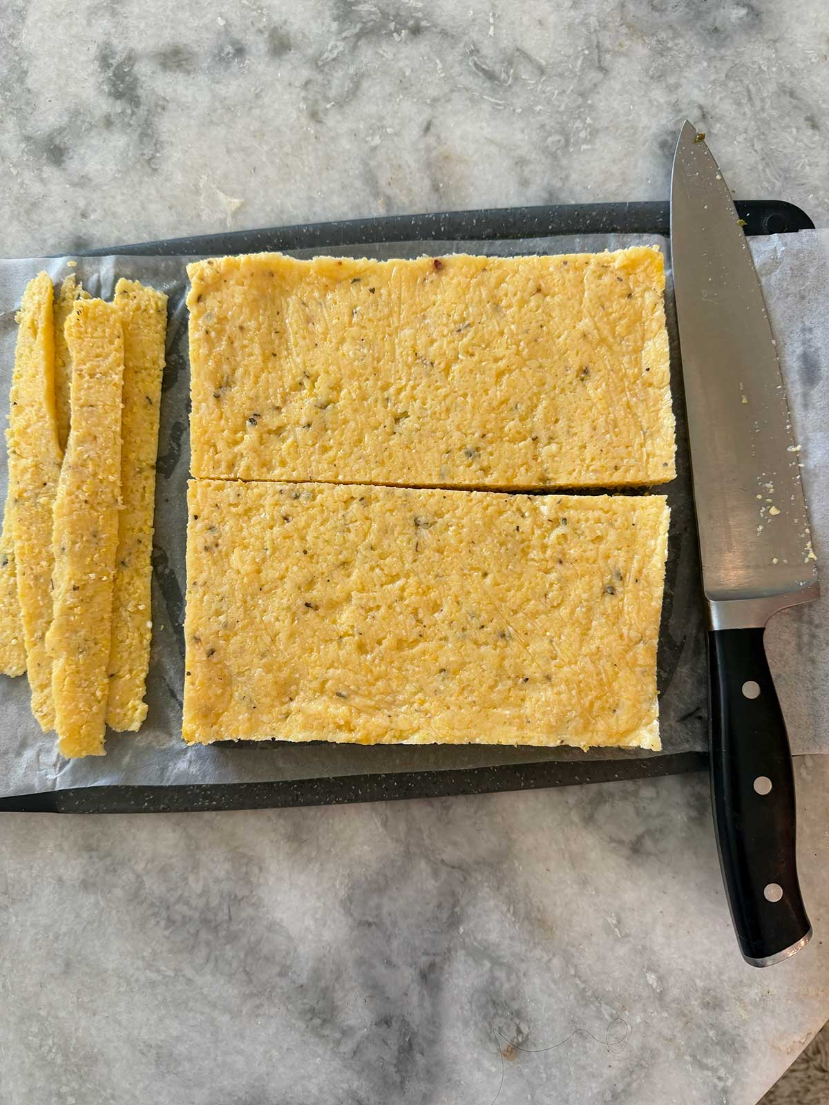 A large knife rests beside a rectangular slab of polenta on parchment paper; several thin strips have been cut from one side. The scene is set on a gray marble countertop.