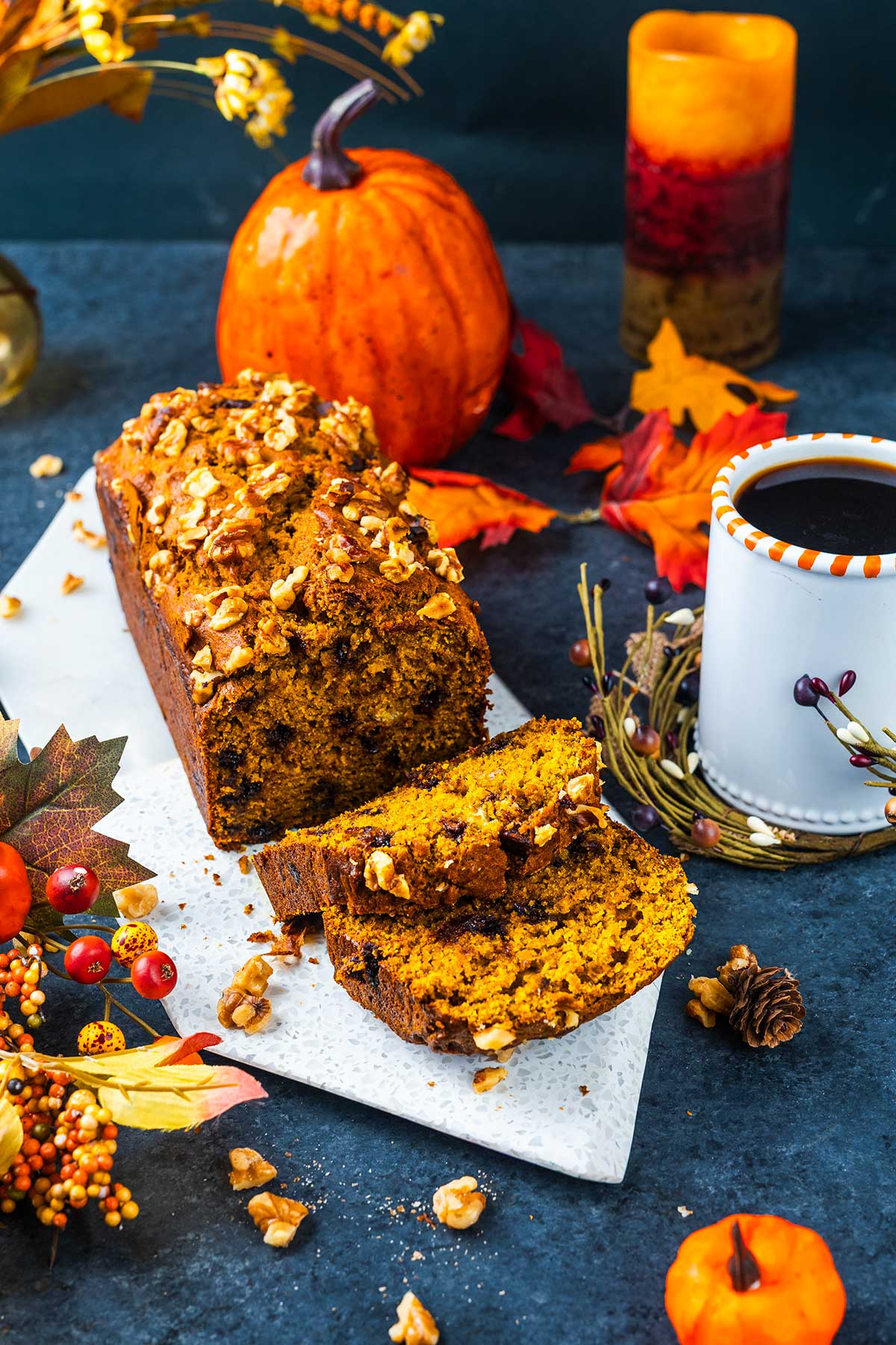 A sliced pumpkin loaf with walnuts sits on a marble board, surrounded by fall décor, pumpkins, a cup of coffee, and colorful autumn leaves. The scene reflects a cozy autumn atmosphere.