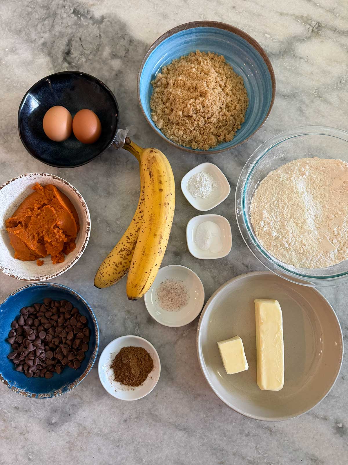 Ingredients on a marble countertop include eggs, brown sugar, flour, bananas, pumpkin puree, butter, chocolate chips, baking soda, baking powder, salt, cinnamon, and nutmeg in various bowls and dishes.