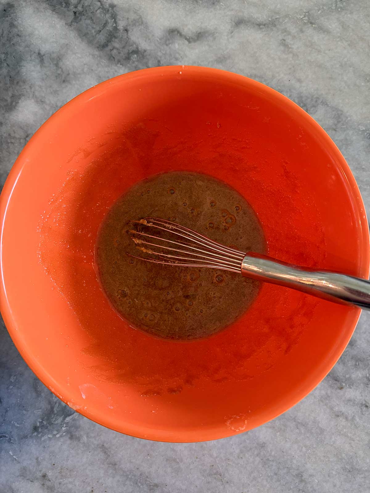 A metal whisk rests in a bright orange mixing bowl containing a brown liquid mixture, set on a gray marble countertop.
