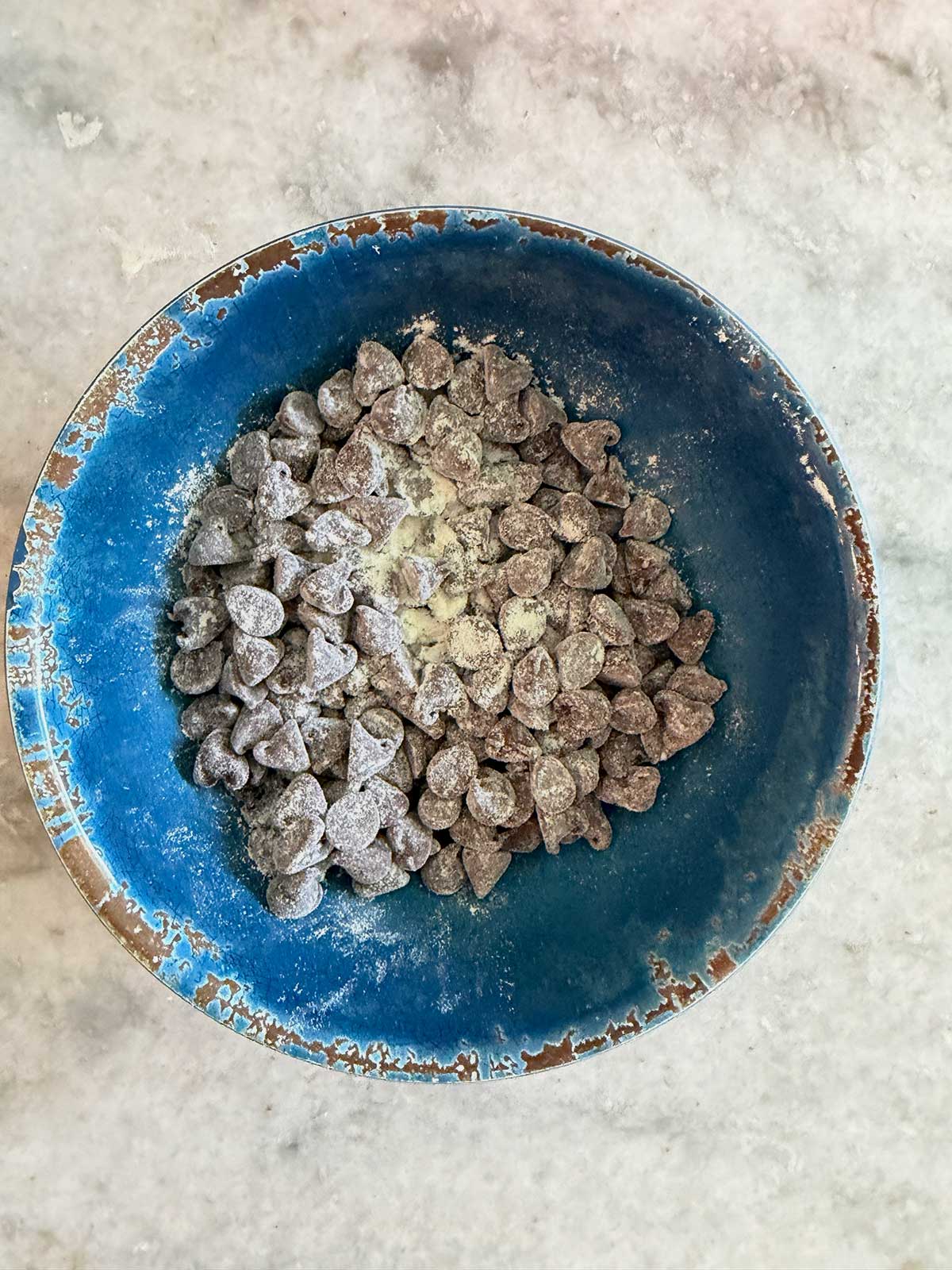 A blue bowl filled with chocolate chips dusted with flour sits on a marble surface.