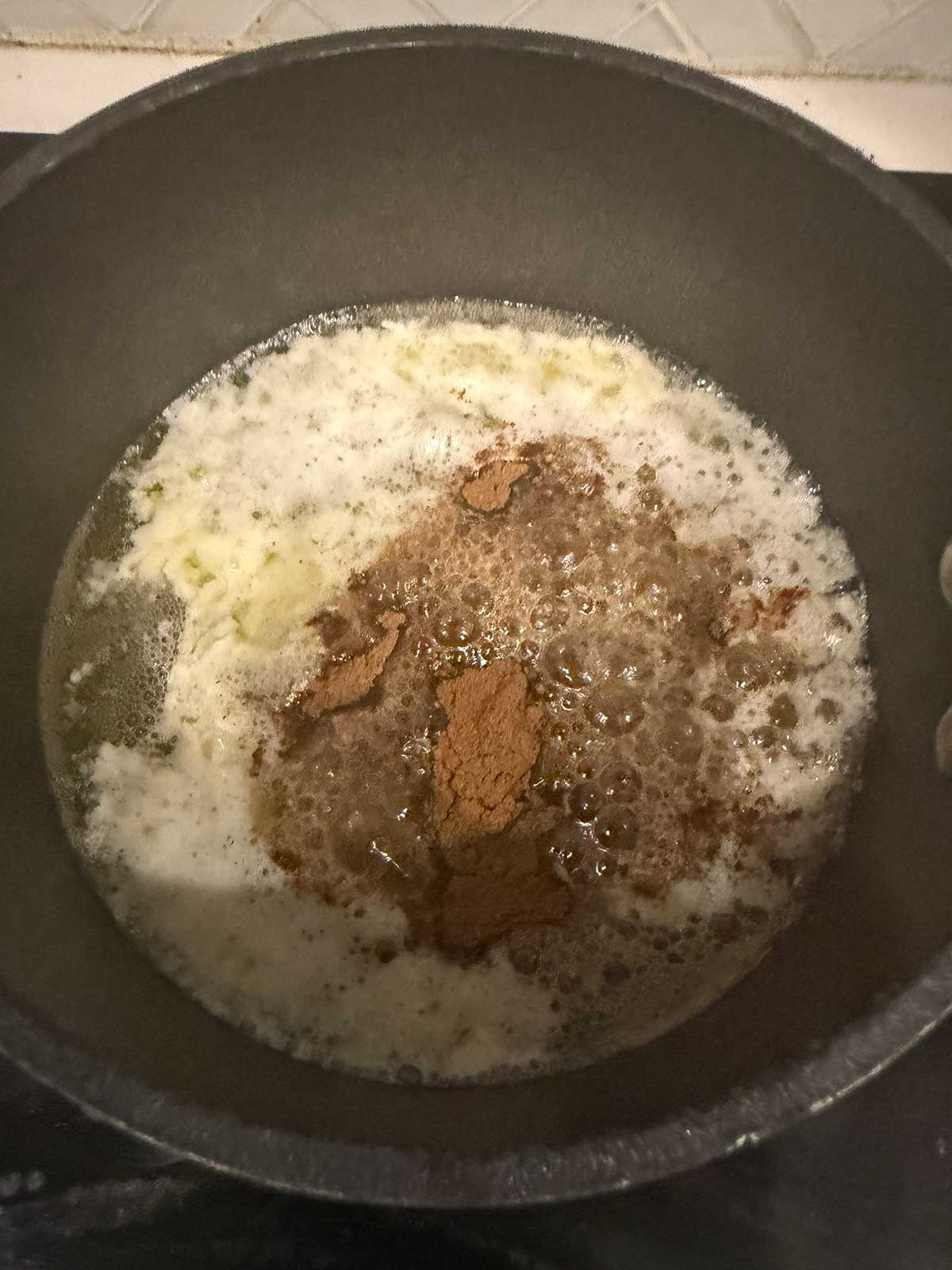 Brown powdered spices are added to melted butter in a black saucepan, with frothy bubbles forming as the mixture cooks on a stovetop. White tile backsplash is partially visible in the background.