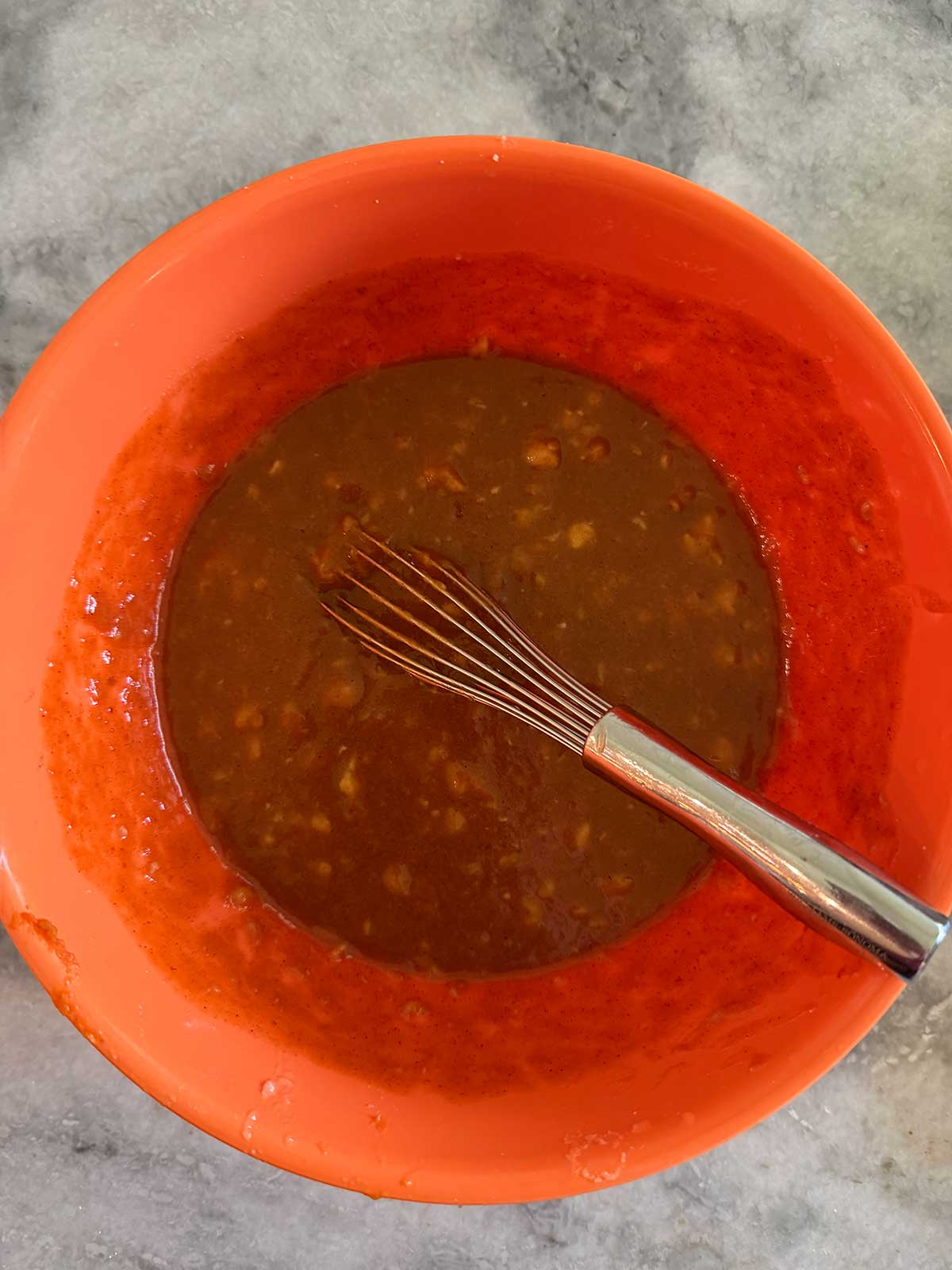 An orange mixing bowl with chocolate batter and a metal whisk sits on a gray countertop.