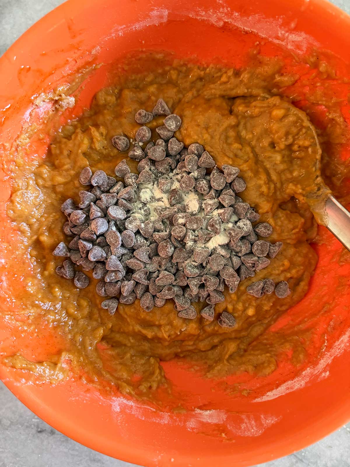 A red mixing bowl with pumpkin bread batter and a pile of chocolate chips in the center, ready to be mixed in with a metal spoon.