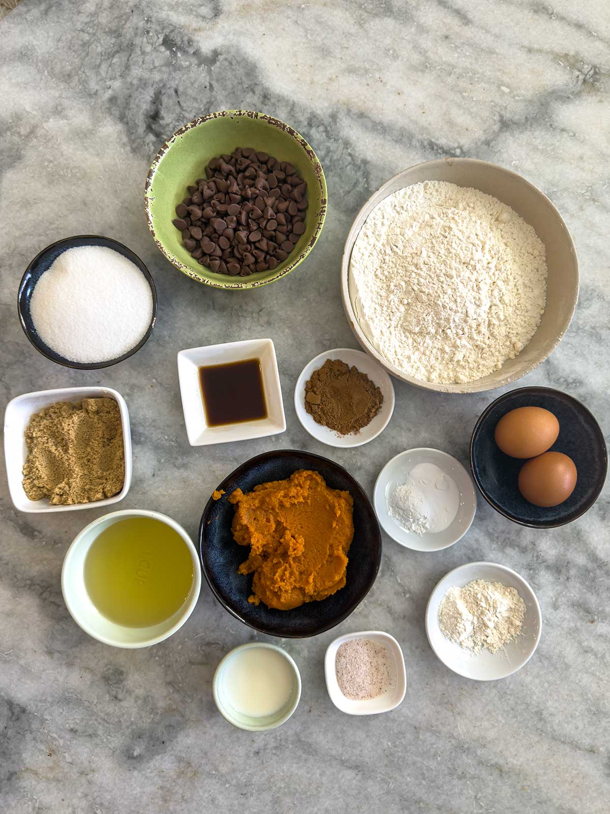 Overhead view of various baking ingredients in bowls on a marble surface, including flour, sugar, chocolate chips, brown sugar, eggs, pumpkin puree, oil, cocoa powder, vanilla, spices, milk, and baking powder.