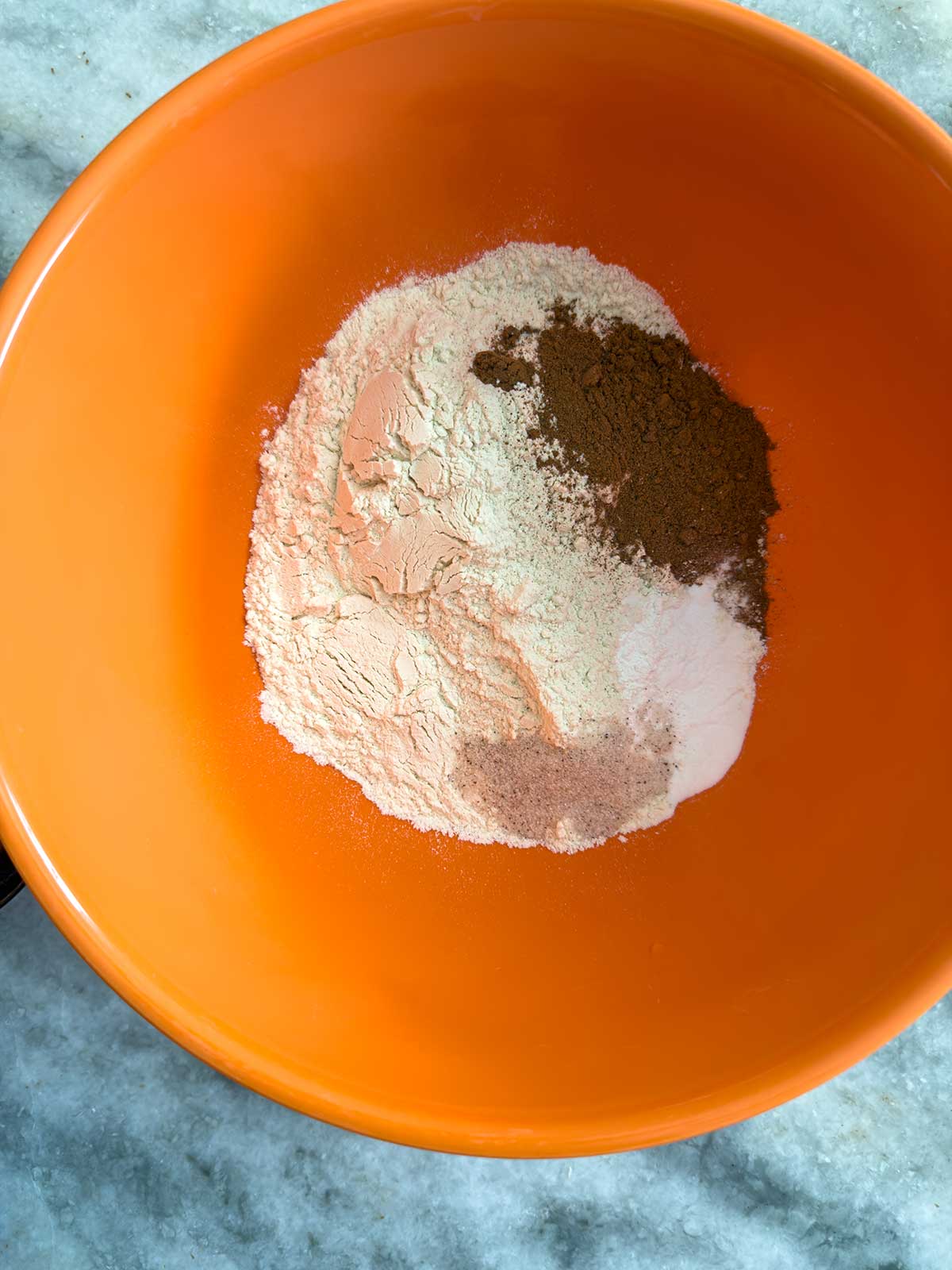 An orange mixing bowl containing piles of dry baking ingredients, including flour, ground cinnamon, and other spices, placed on a light-colored marble countertop.