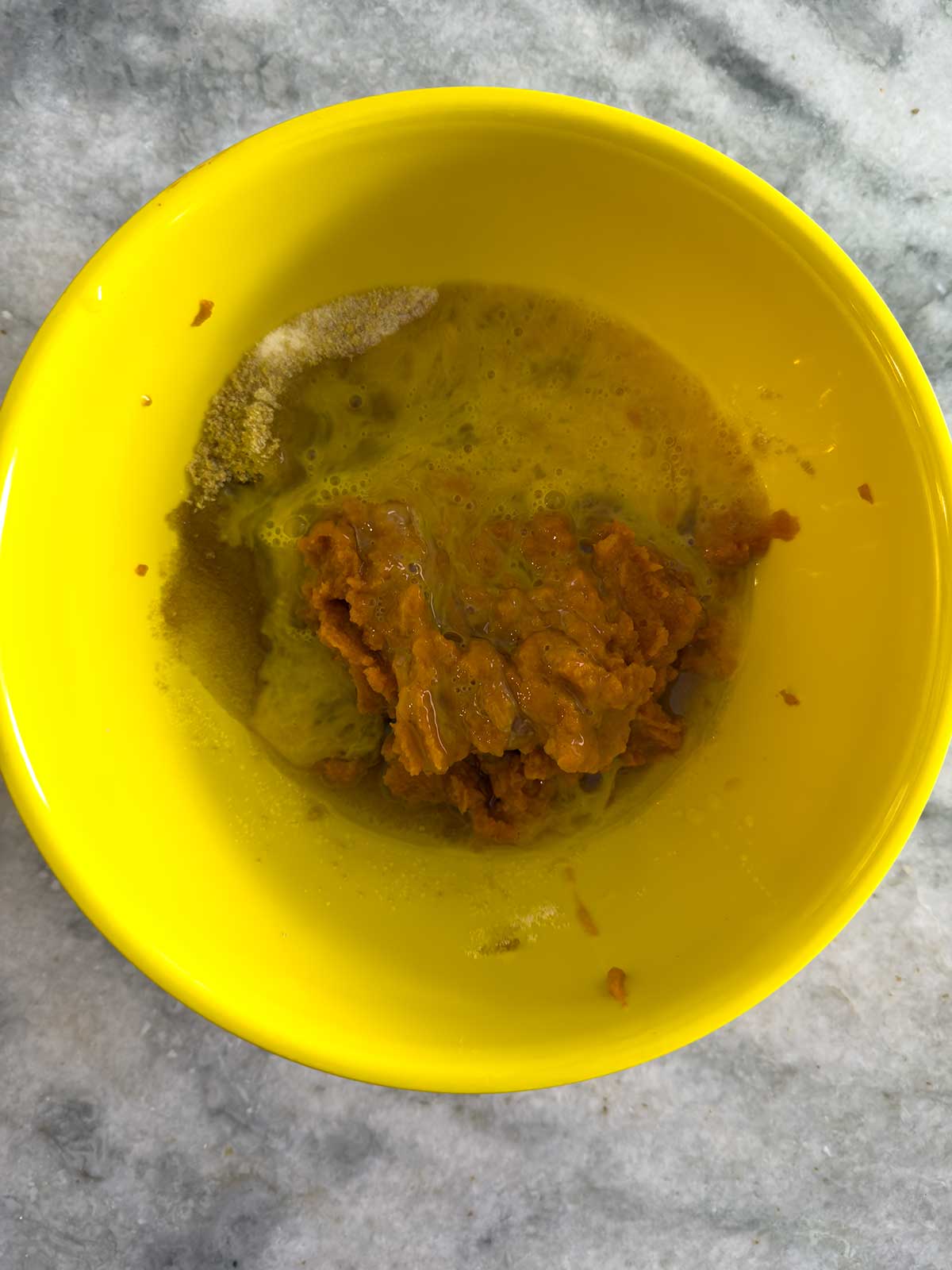 A yellow bowl contains a mixture of orange pumpkin puree, a cracked egg with visible yolk, and some granulated white sugar on a marble countertop.