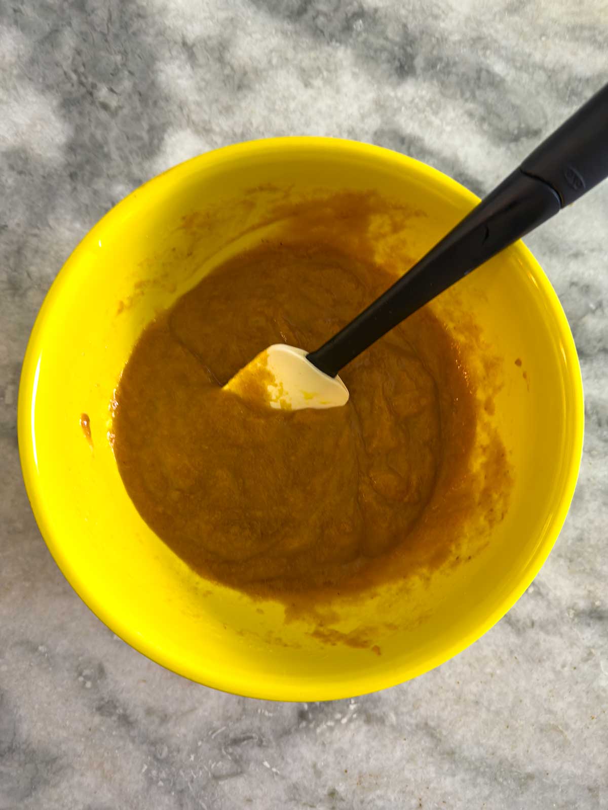 A bright yellow bowl containing a thick orange-brown mixture with a black-handled white spatula resting inside, placed on a gray marble surface.