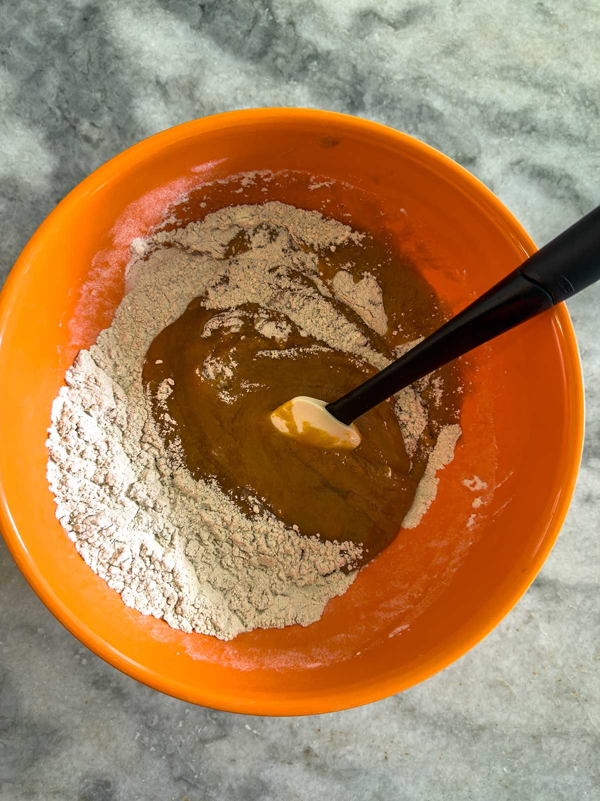 An orange mixing bowl on a gray countertop contains flour and a brown liquid being mixed with a black and white spatula.