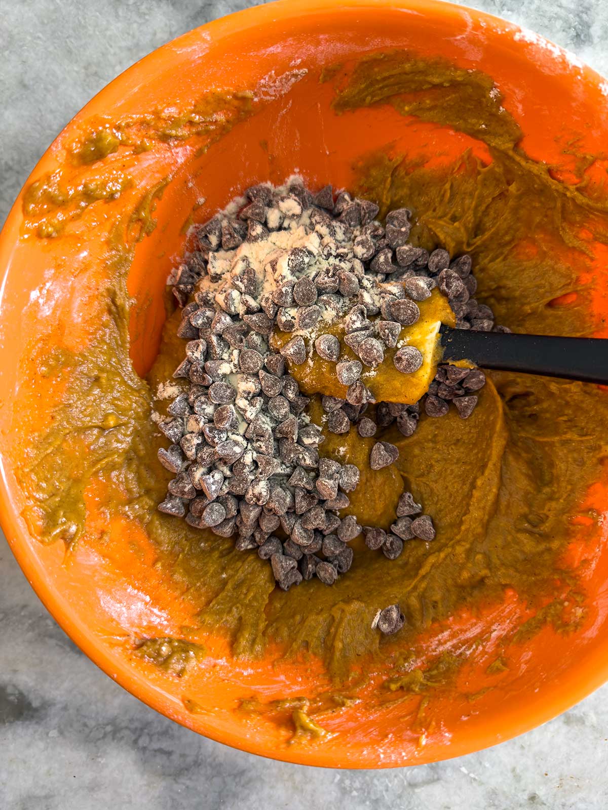 An orange mixing bowl with chocolate chip cookie dough and a pile of chocolate chips being stirred in with a black spatula, on a gray countertop.
