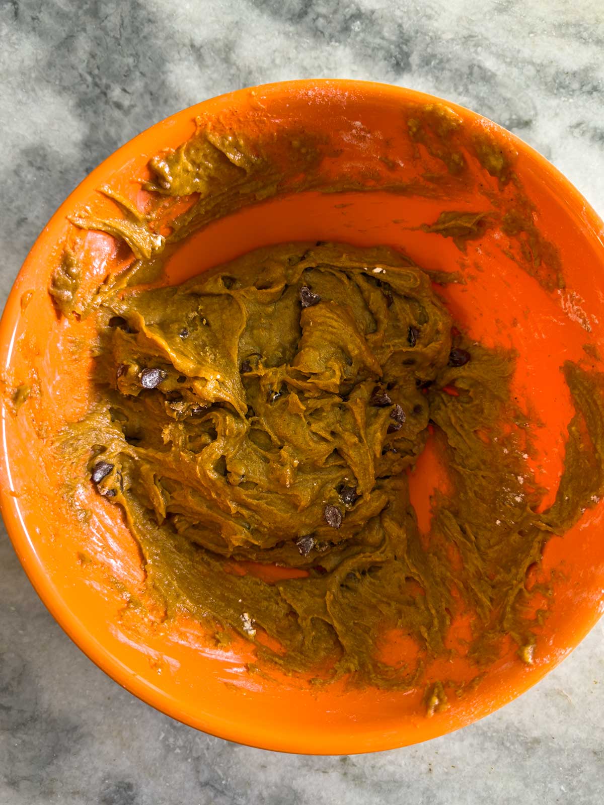 An orange mixing bowl filled with cookie dough mixed with chocolate chips, placed on a gray marble countertop.